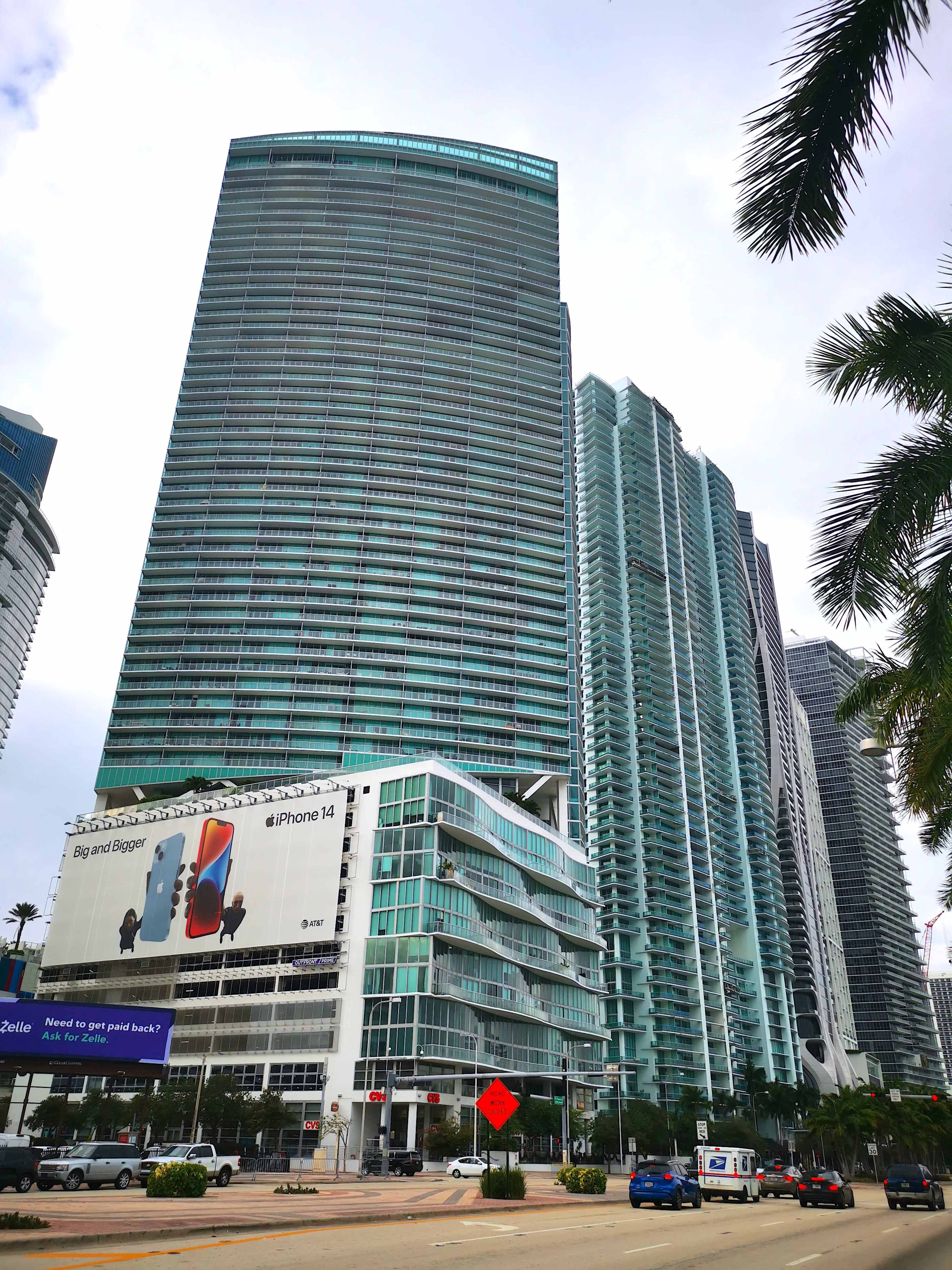 A towering modern skyscraper stands alongside a busy road, with an advertisement for the iPhone 14 visible on a building's facade.