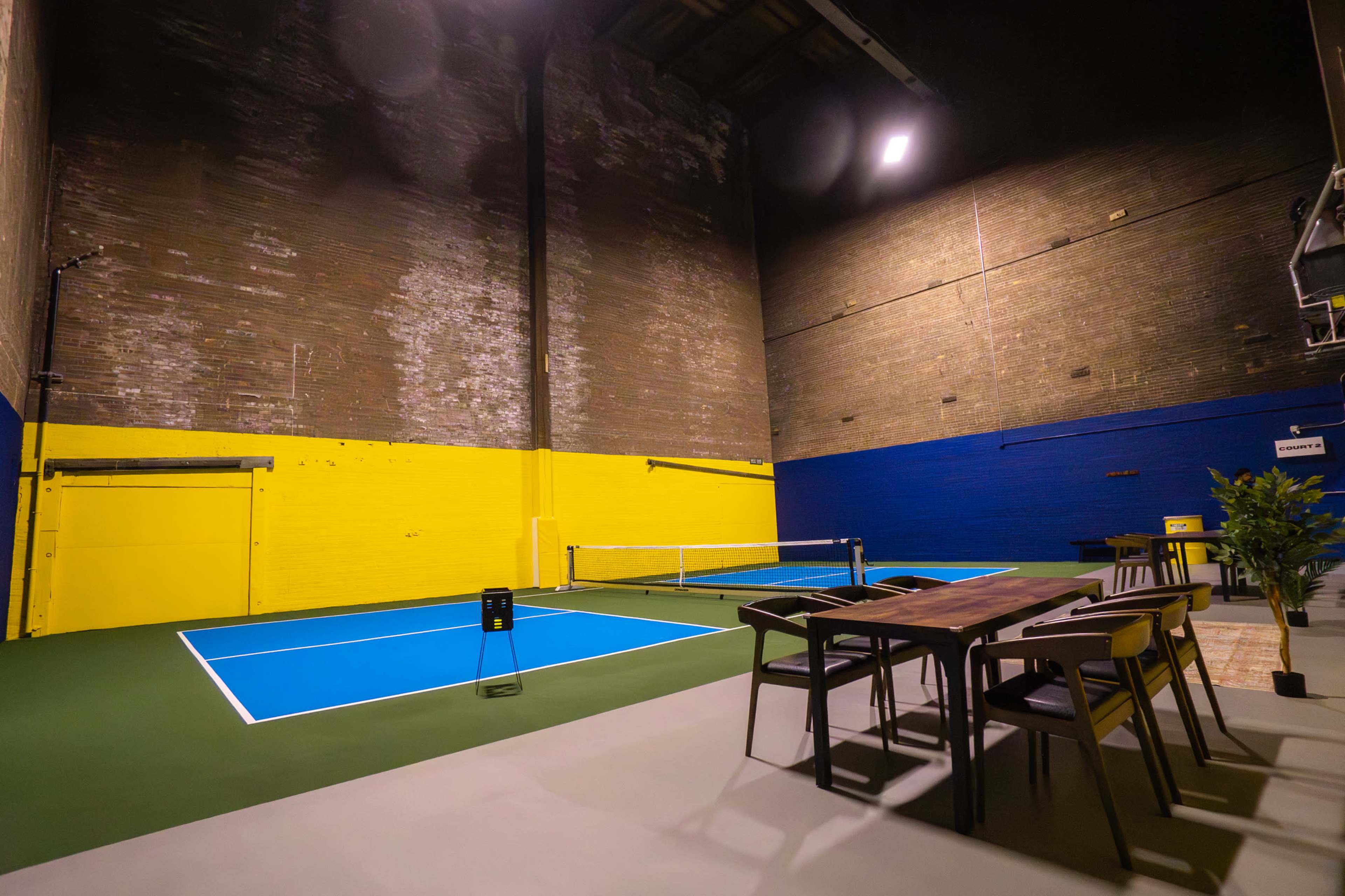 The image shows an indoor tennis court with blue playing surfaces and yellow and blue painted brick walls, along with a dining area featuring a table and chairs nearby.