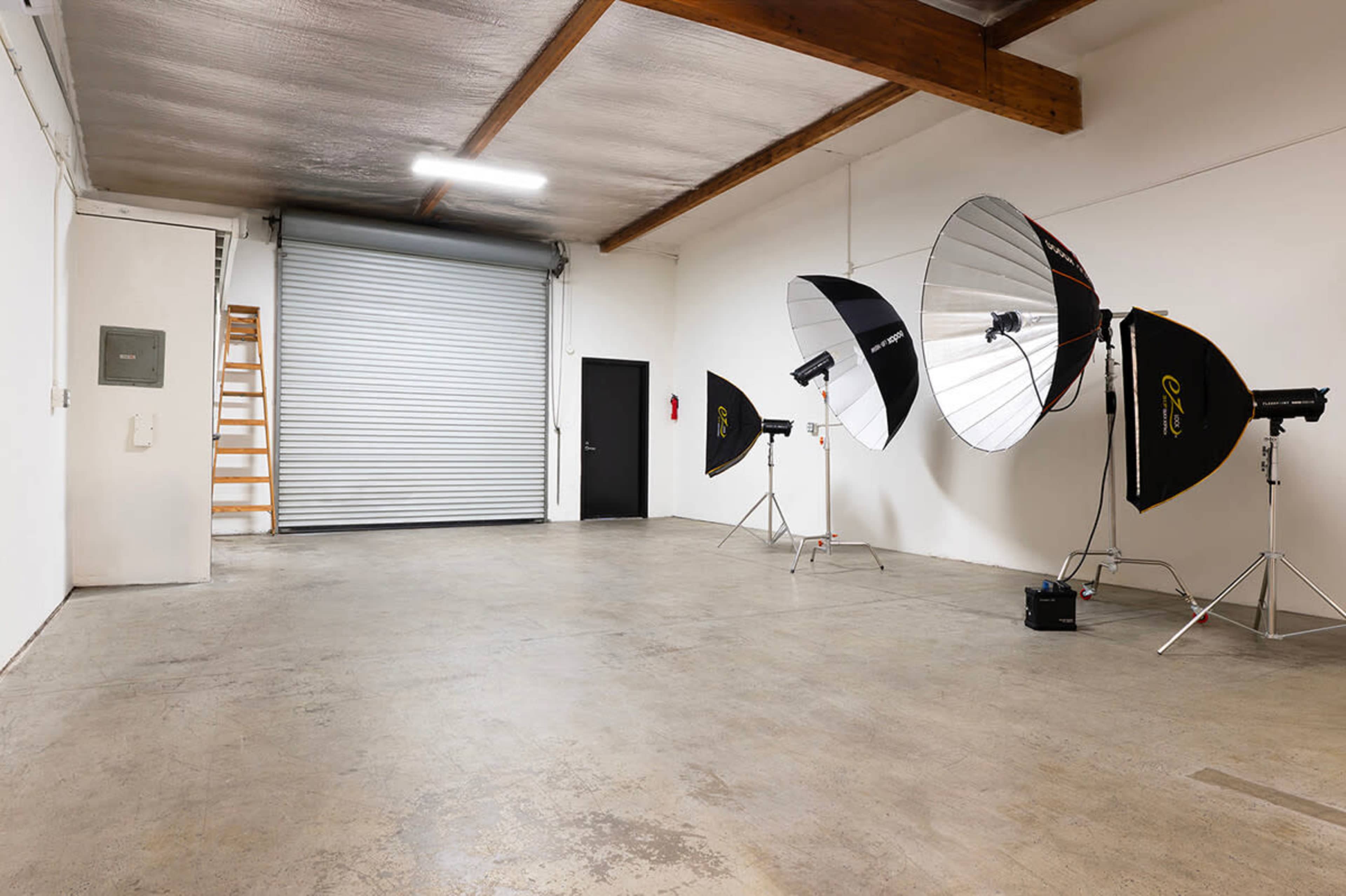 The image shows a large, empty studio space with concrete flooring and three photography light setups, along with a closed rolling door and a ladder against the wall.