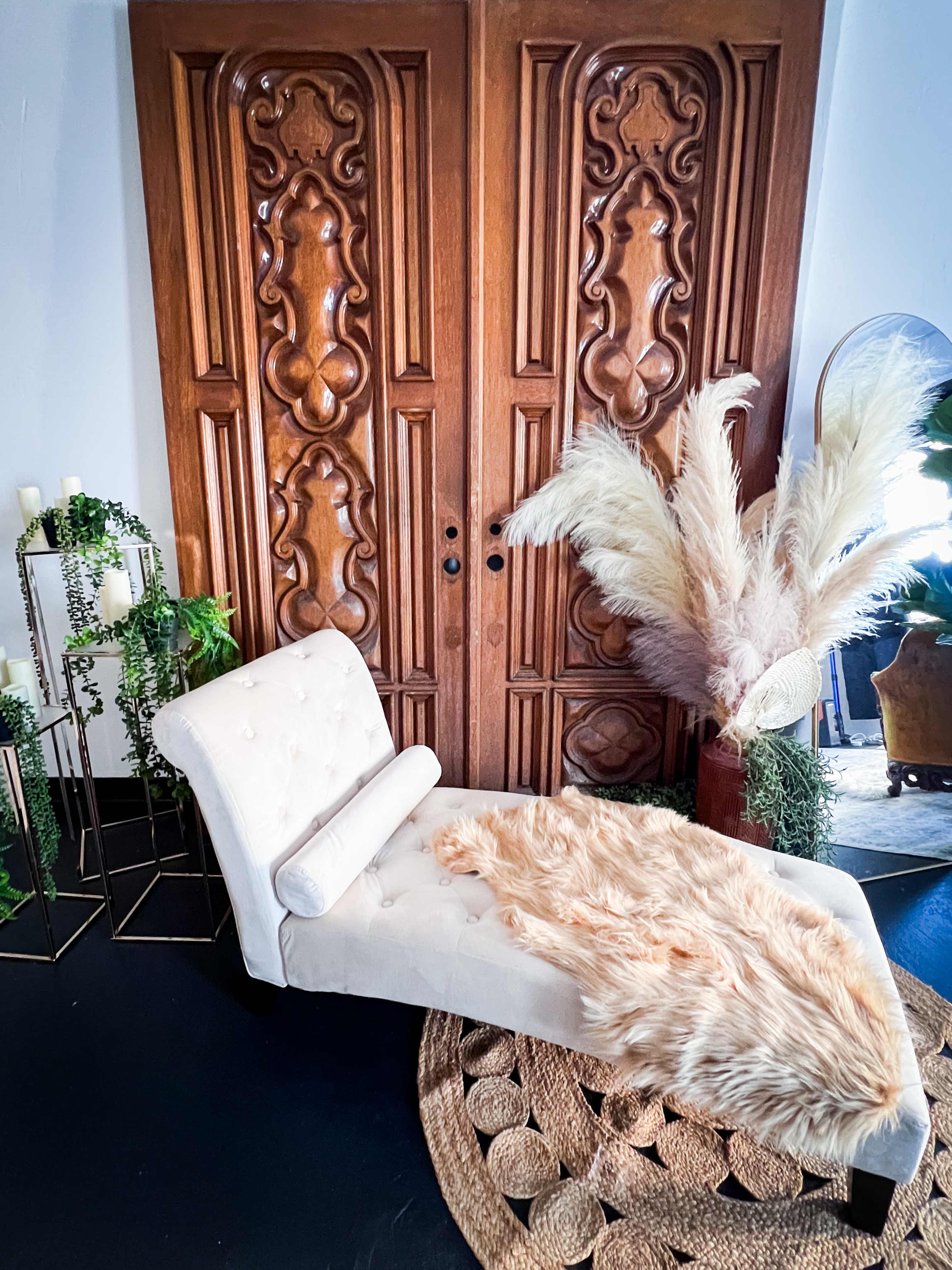 A white chaise lounge with a faux fur throw is positioned in front of an intricately carved wooden door, surrounded by decorative plants and a mirror.
