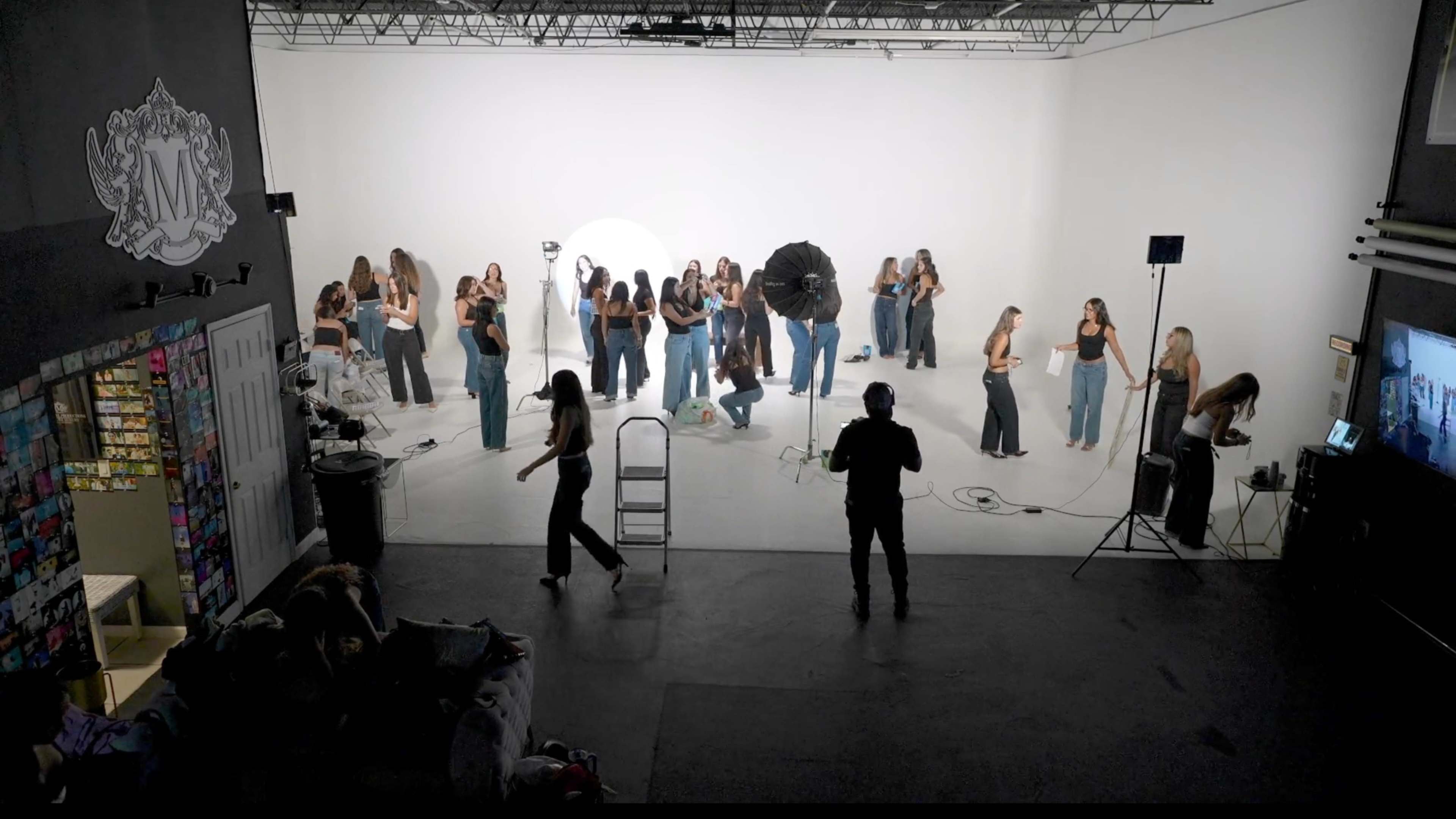 A photography studio is filled with a group of models and crew members preparing for a shoot in front of a seamless white backdrop.