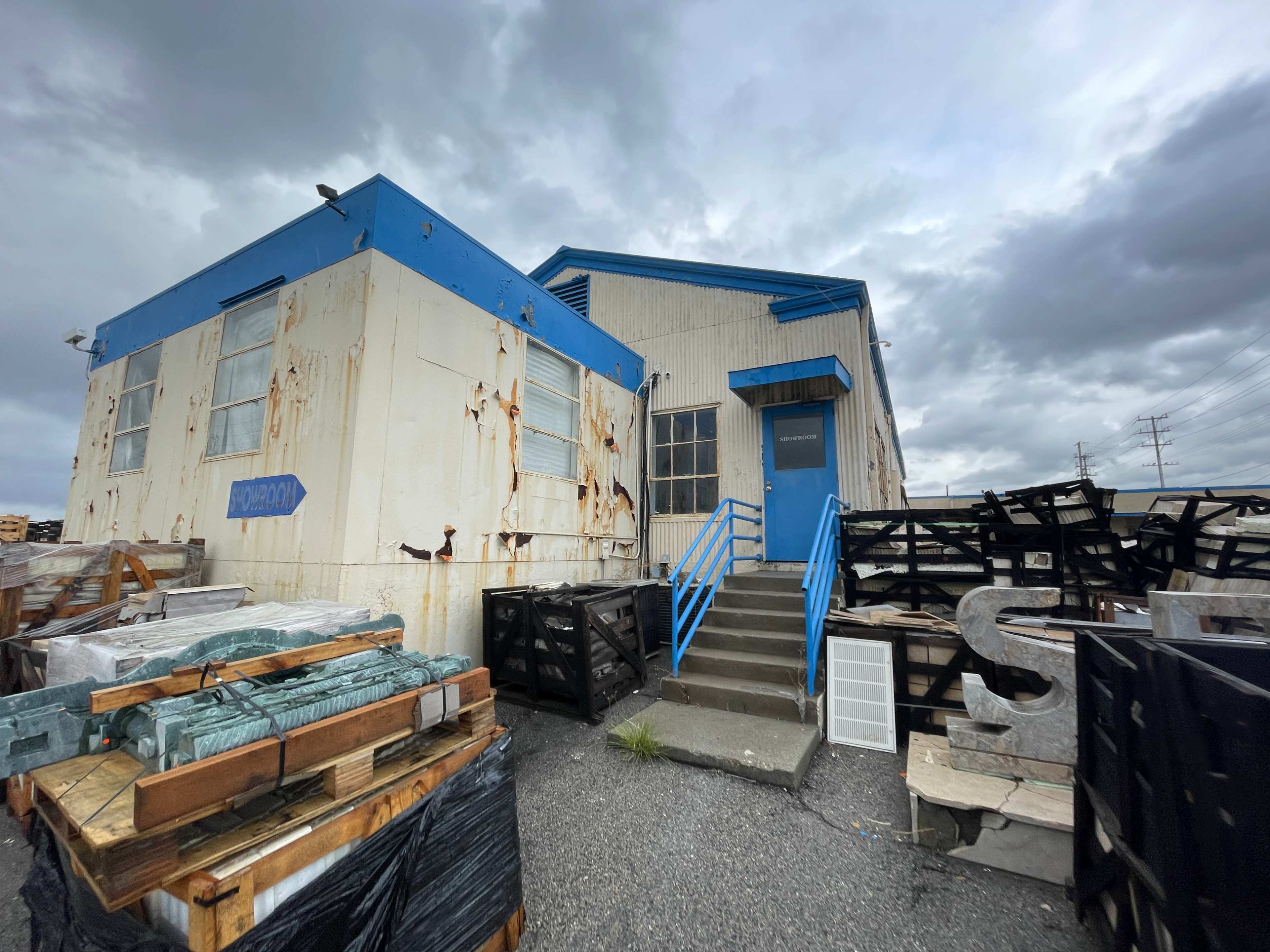A weathered building with a blue roof and trim is situated next to stacks of shipping crates and black pallets under a cloudy sky.