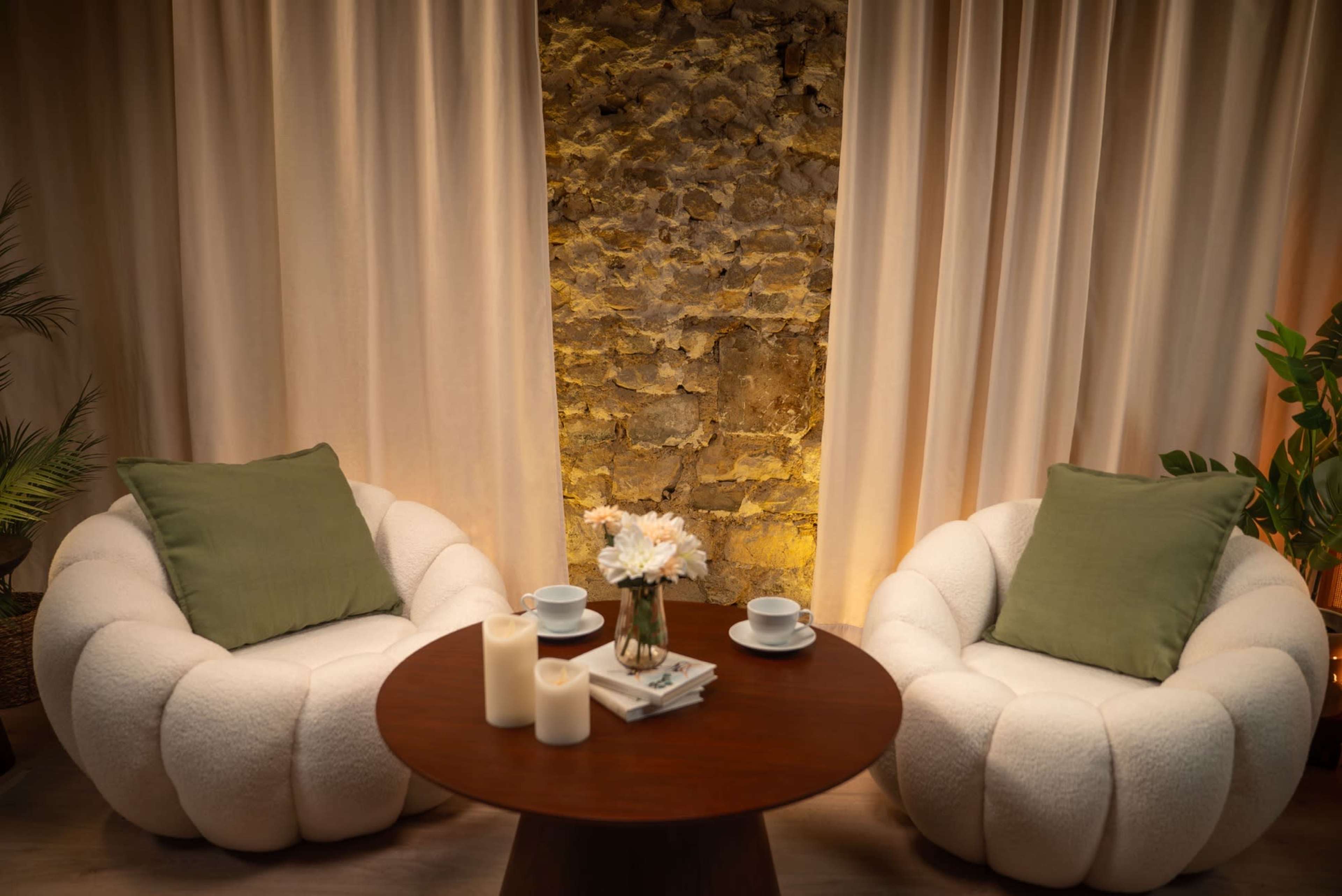 The image shows two plush, round chairs with green cushions facing a wooden table adorned with a vase of flowers and three candles, set against a rustic stone wall and fabric curtains.
