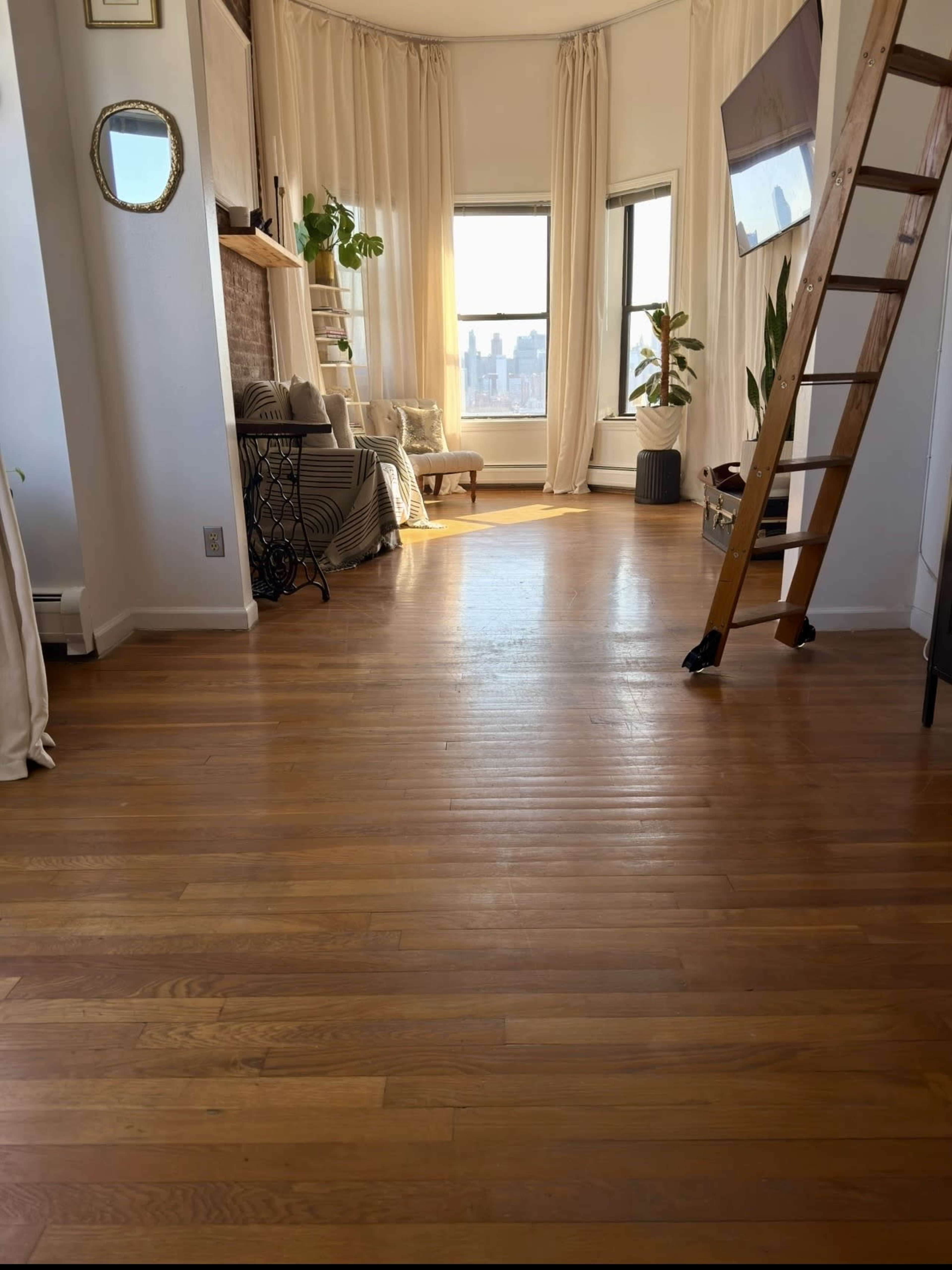 The image shows a spacious, sunlit room with wooden floors, a large window, a ladder against the wall, and a seating area with plants and decor.
