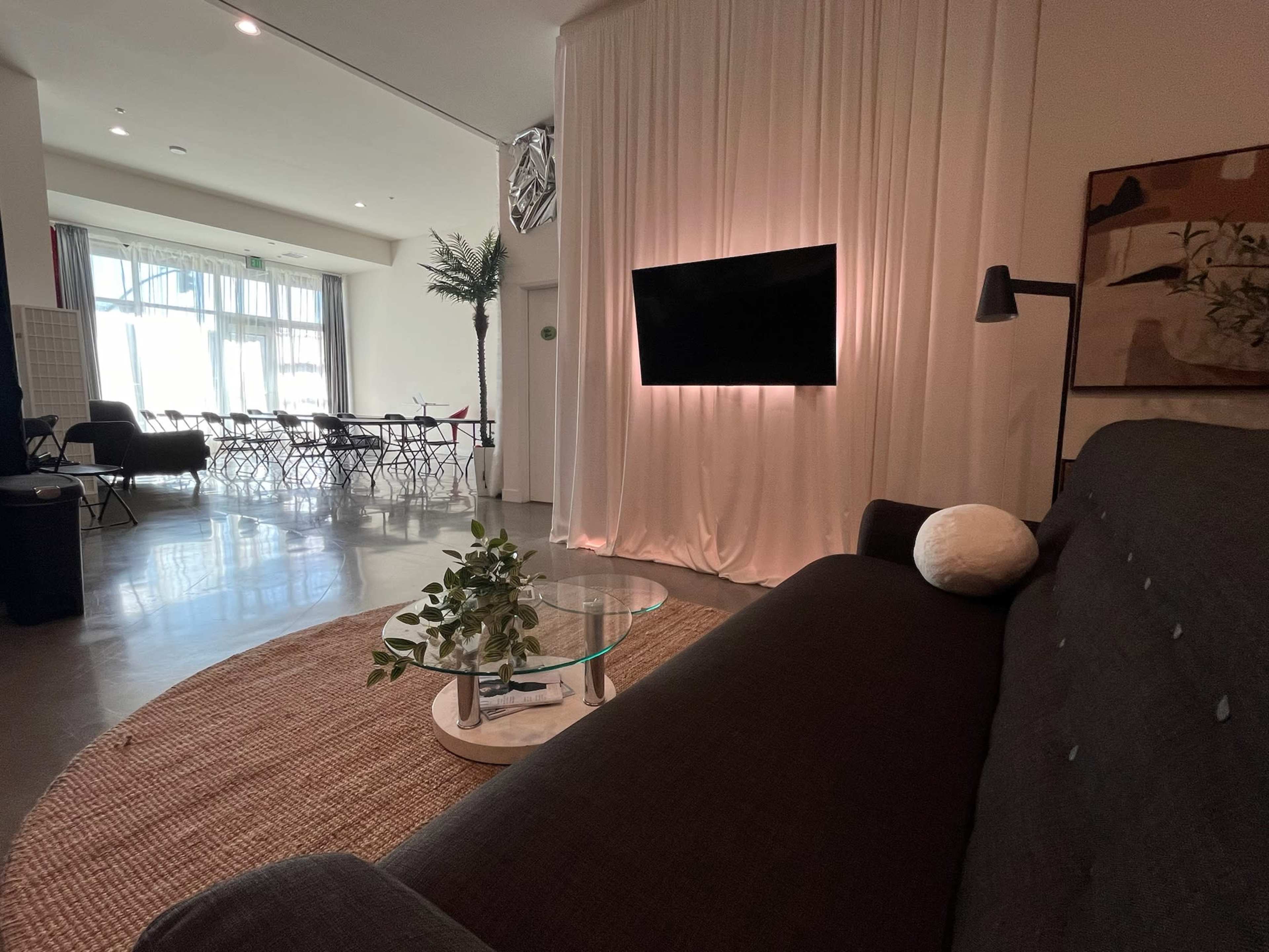 A modern living space featuring a dark sofa, a glass coffee table with a plant, a wall-mounted television, and an area with white drapes in the background.