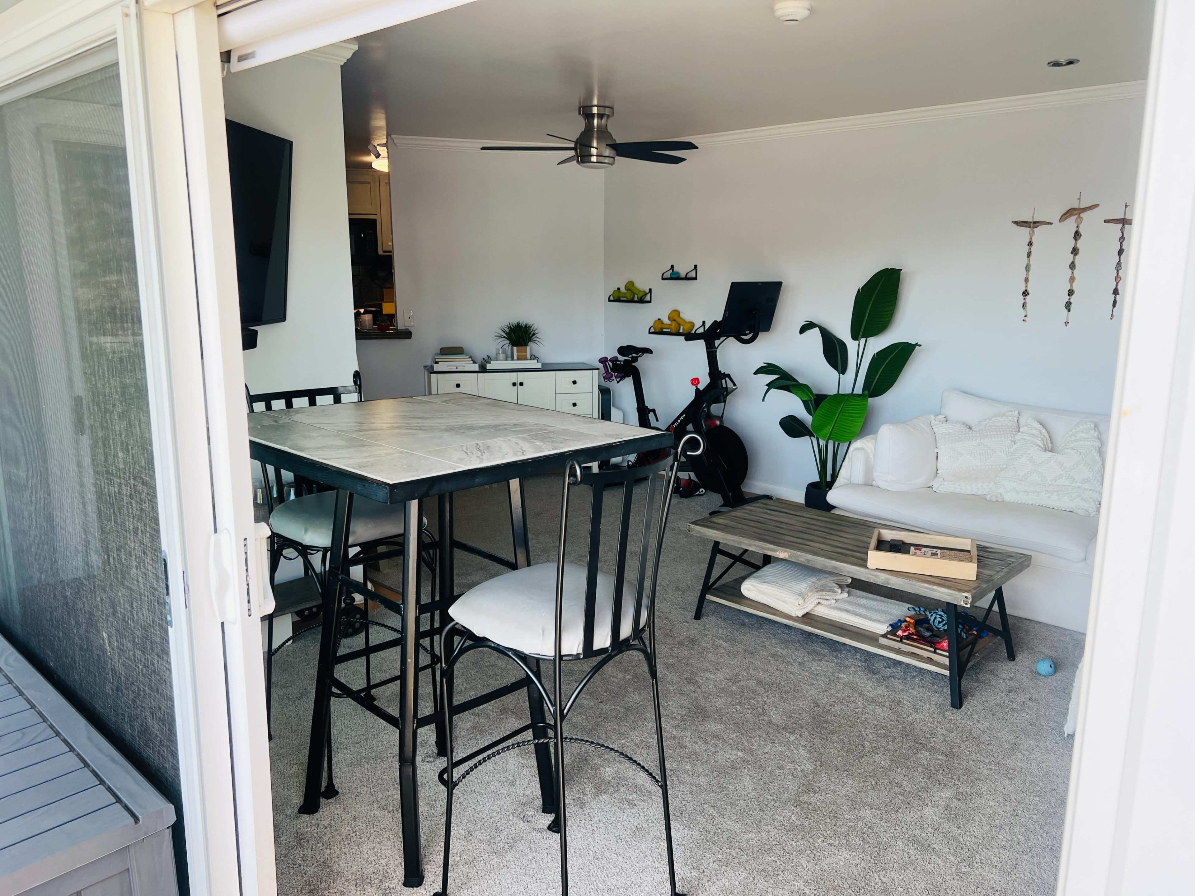 The image shows a living area with a dining table, chairs, a sofa, and exercise equipment, all set on carpeted flooring.