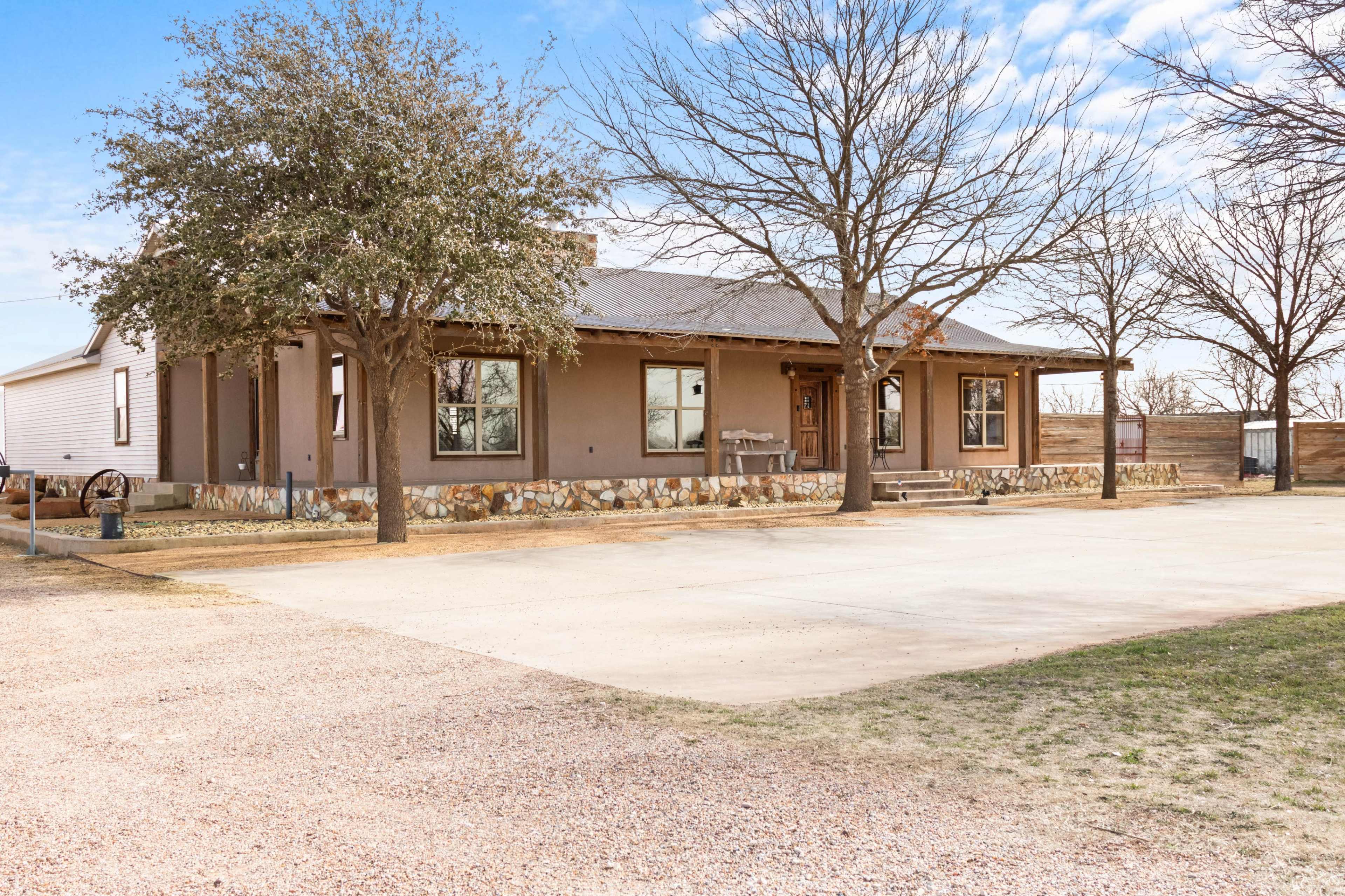 The image shows a one-story house with a stone foundation, surrounded by bare trees, set on a gravel and paved area.