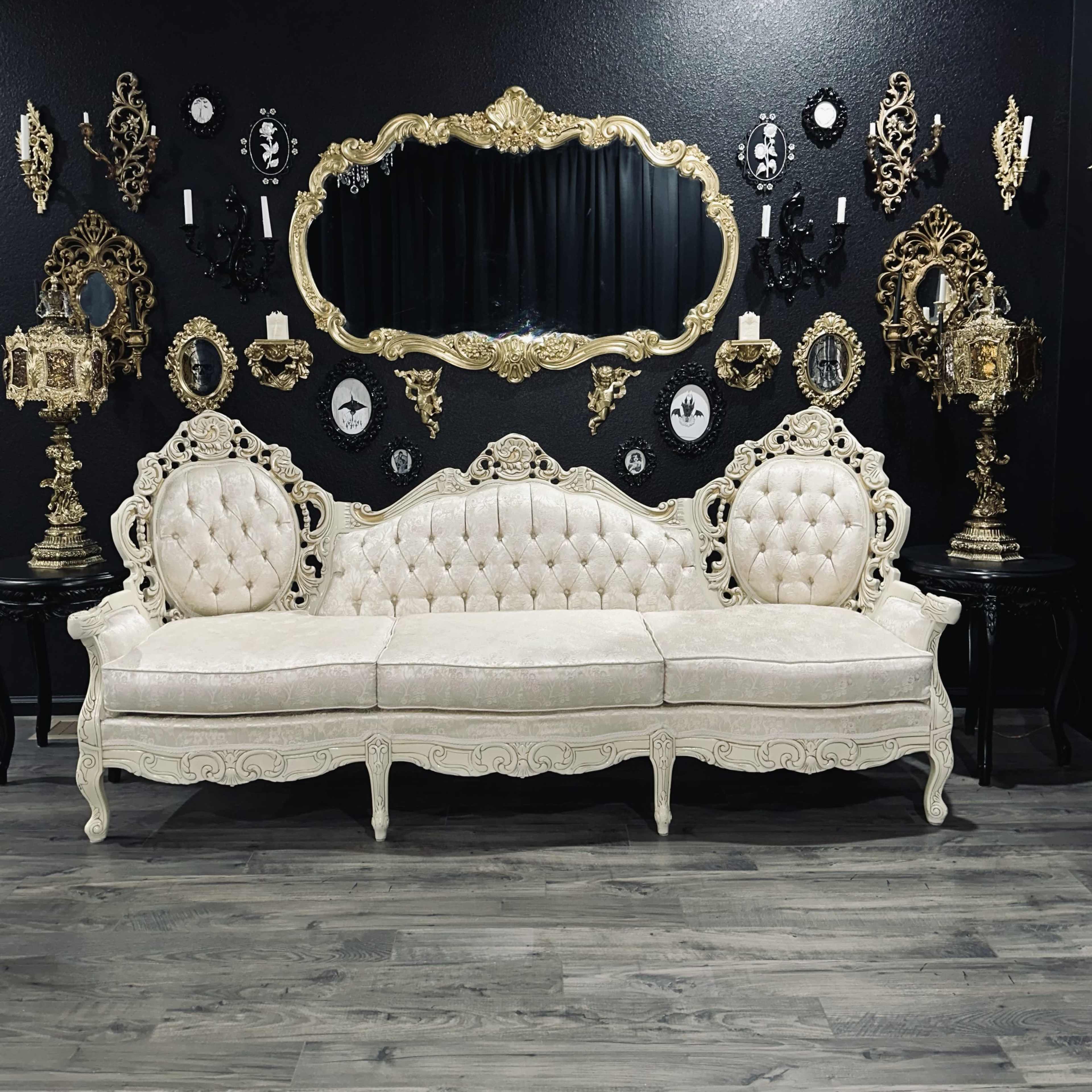 A vintage ornate white sofa is positioned in front of a large decorative mirror, surrounded by framed artwork and antique wall decorations.