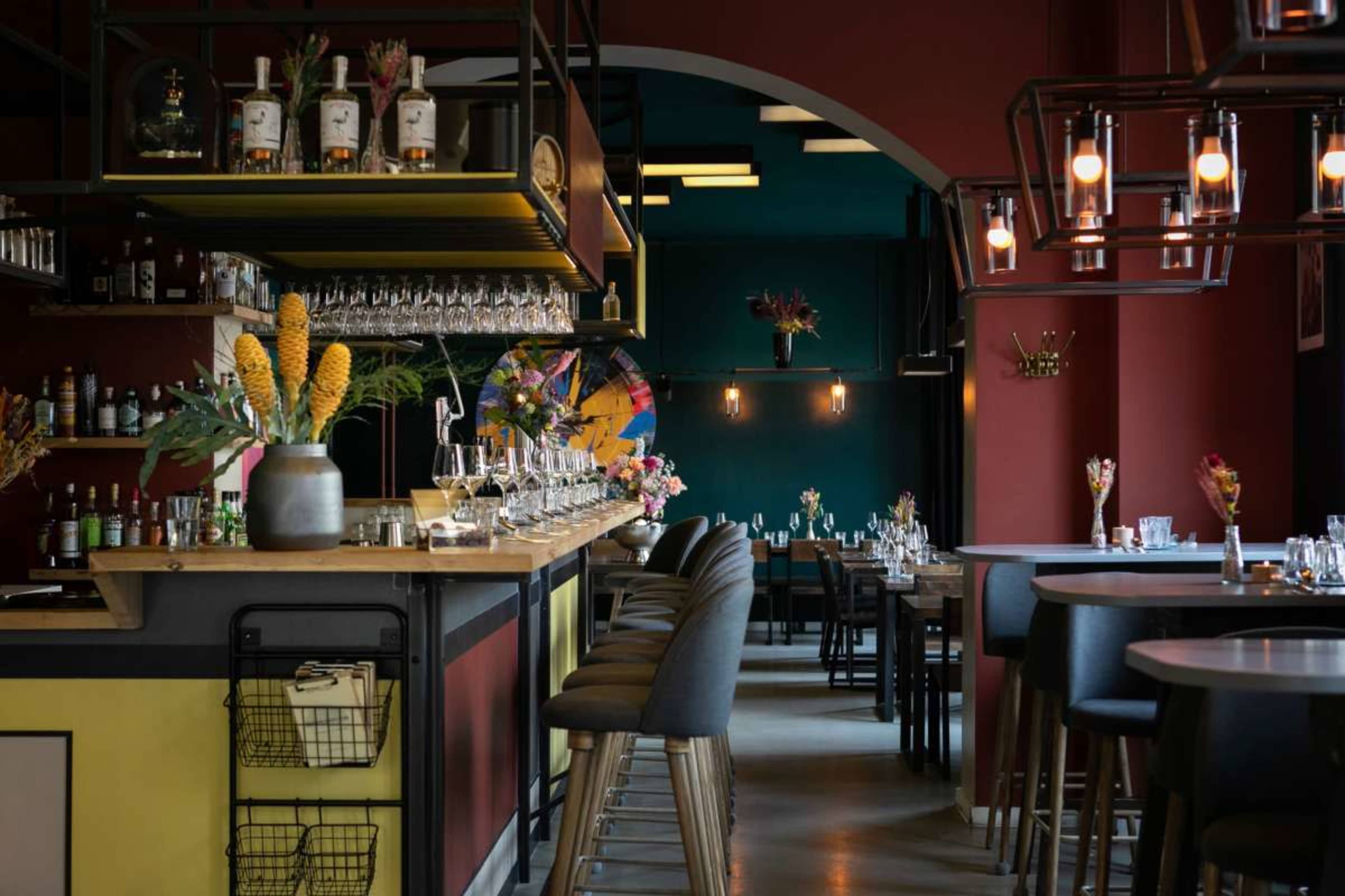 The image shows a modern restaurant interior featuring a long bar with high stools, colorful walls, and floral decorations arranged on tables.