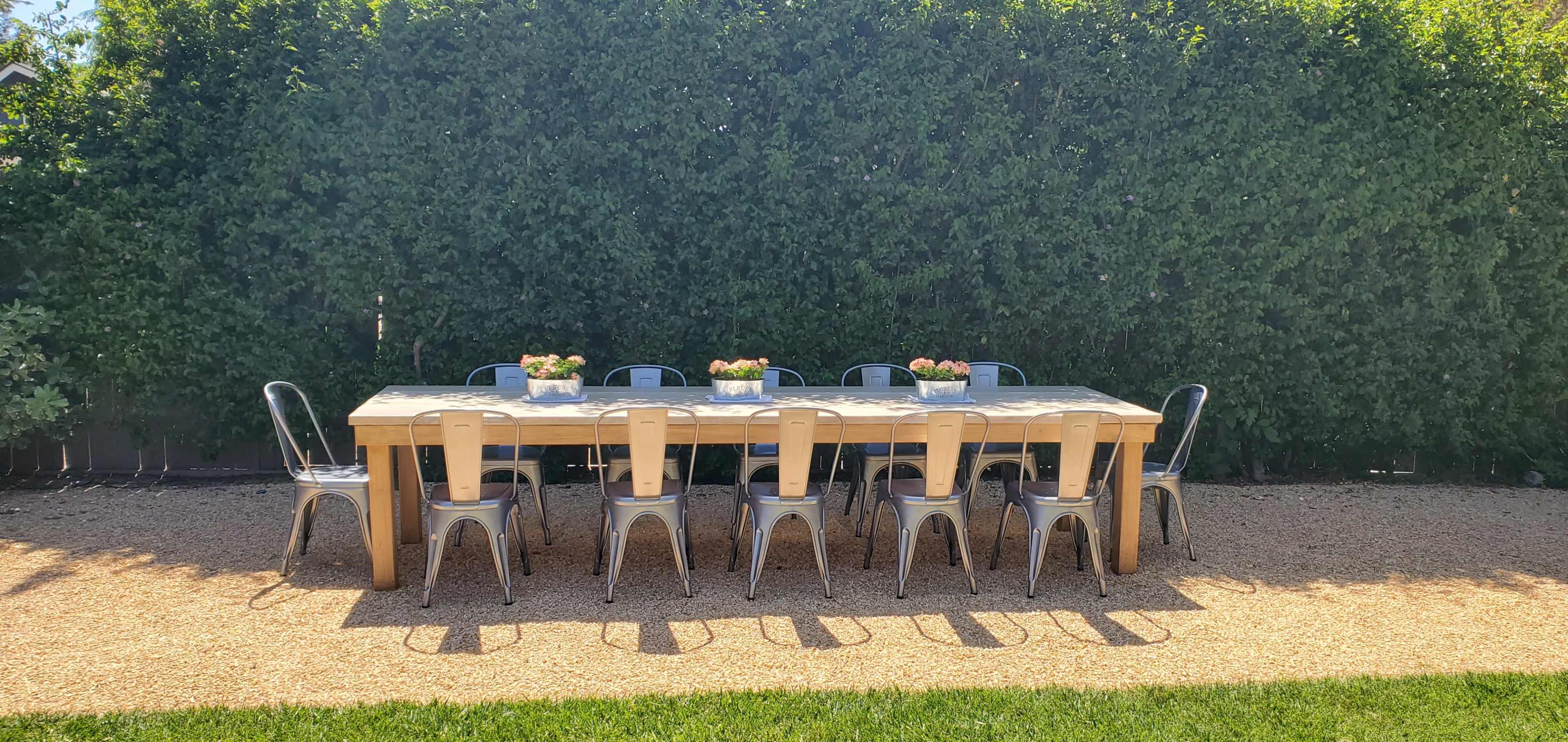 A long wooden dining table with metal chairs is set in a garden surrounded by a dense hedge.