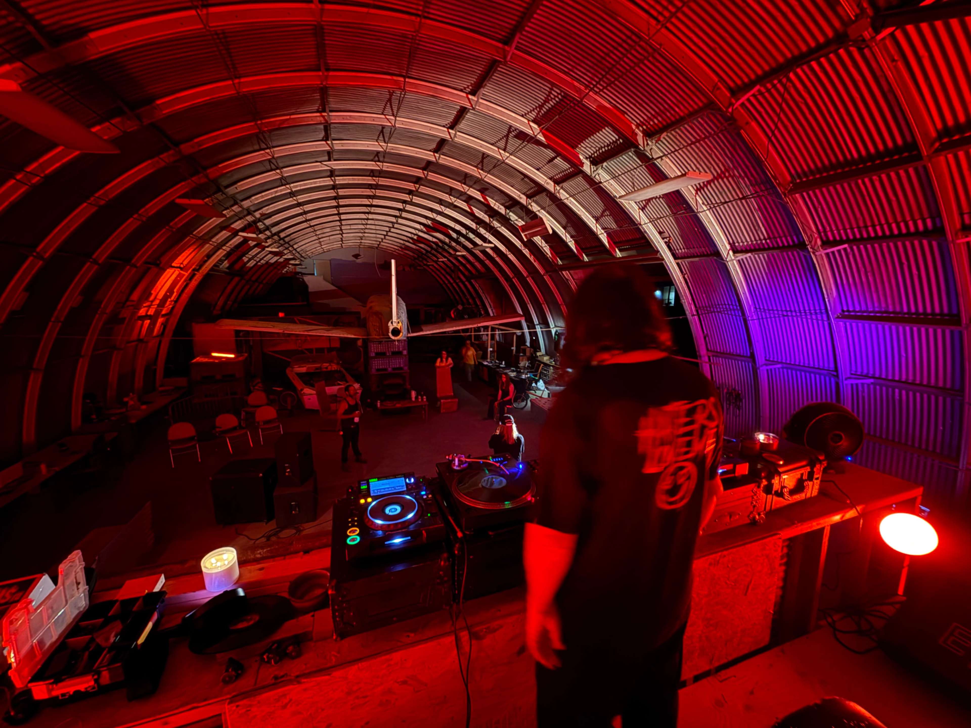 A DJ stands behind turntables in a dimly lit, arched warehouse filled with colorful lights while people dance and socialize.