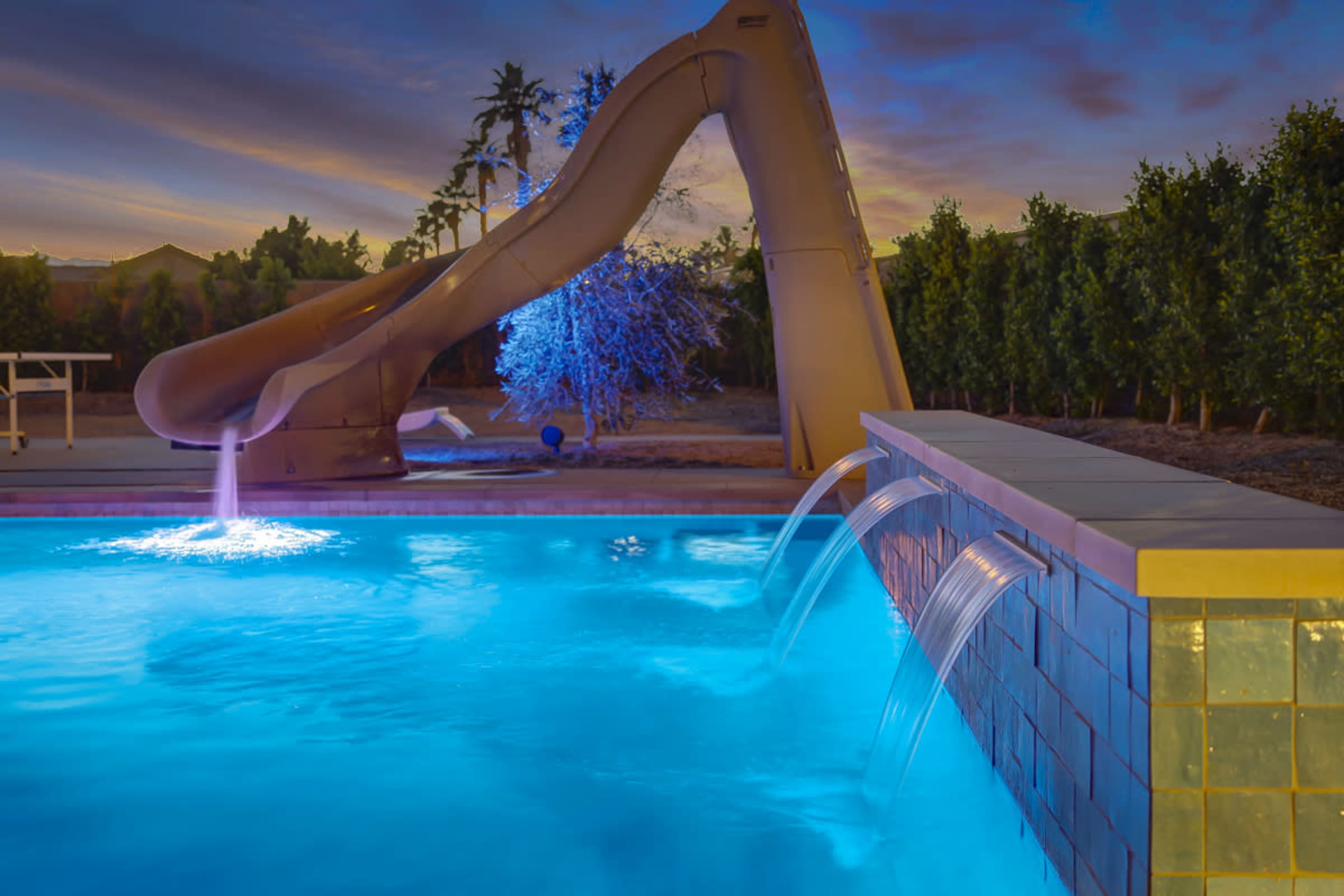 A large water slide stands beside a brightly lit swimming pool with flowing water features.
