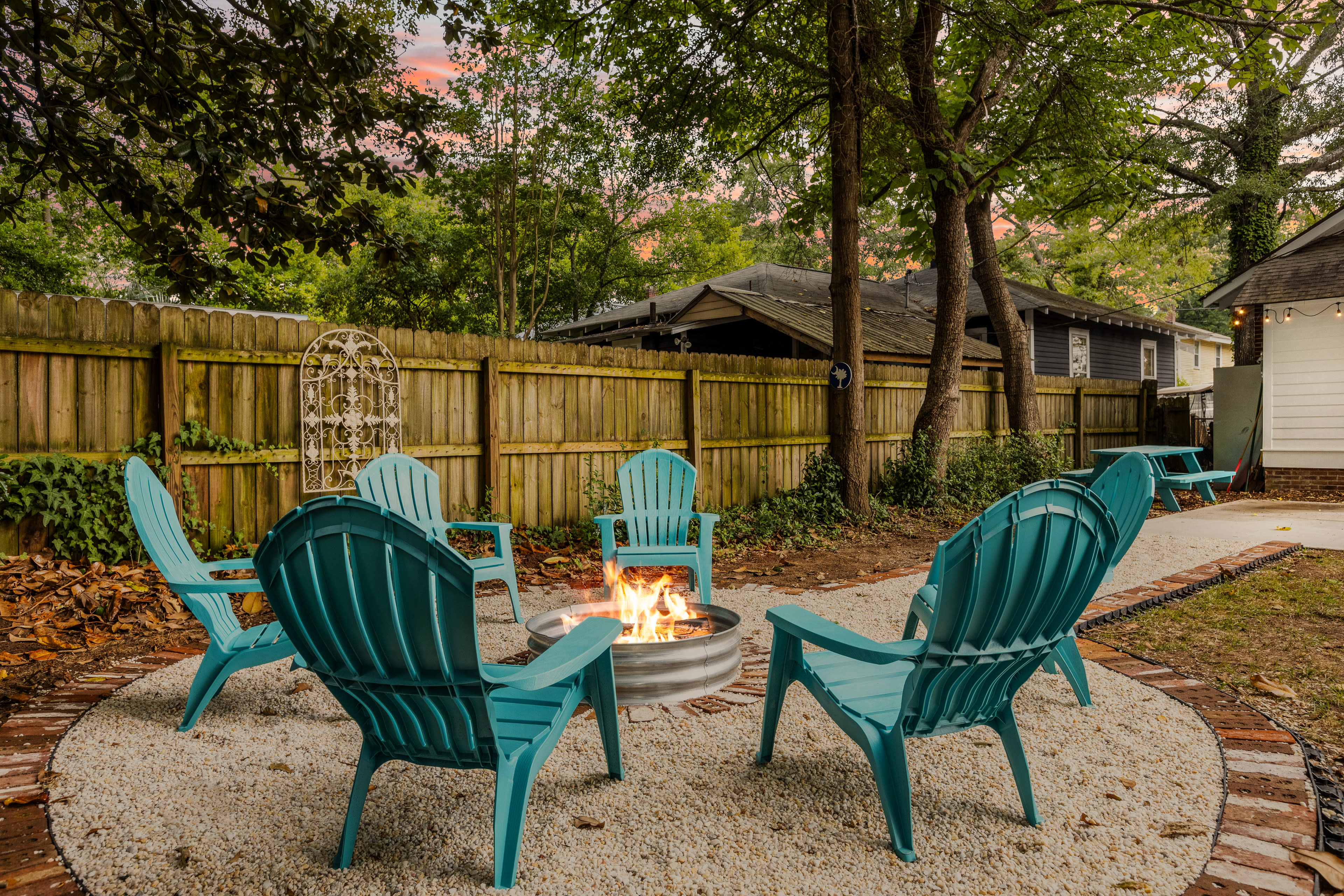 A circular stone patio features five teal chairs arranged around a fire pit, surrounded by a wooden fence and greenery.