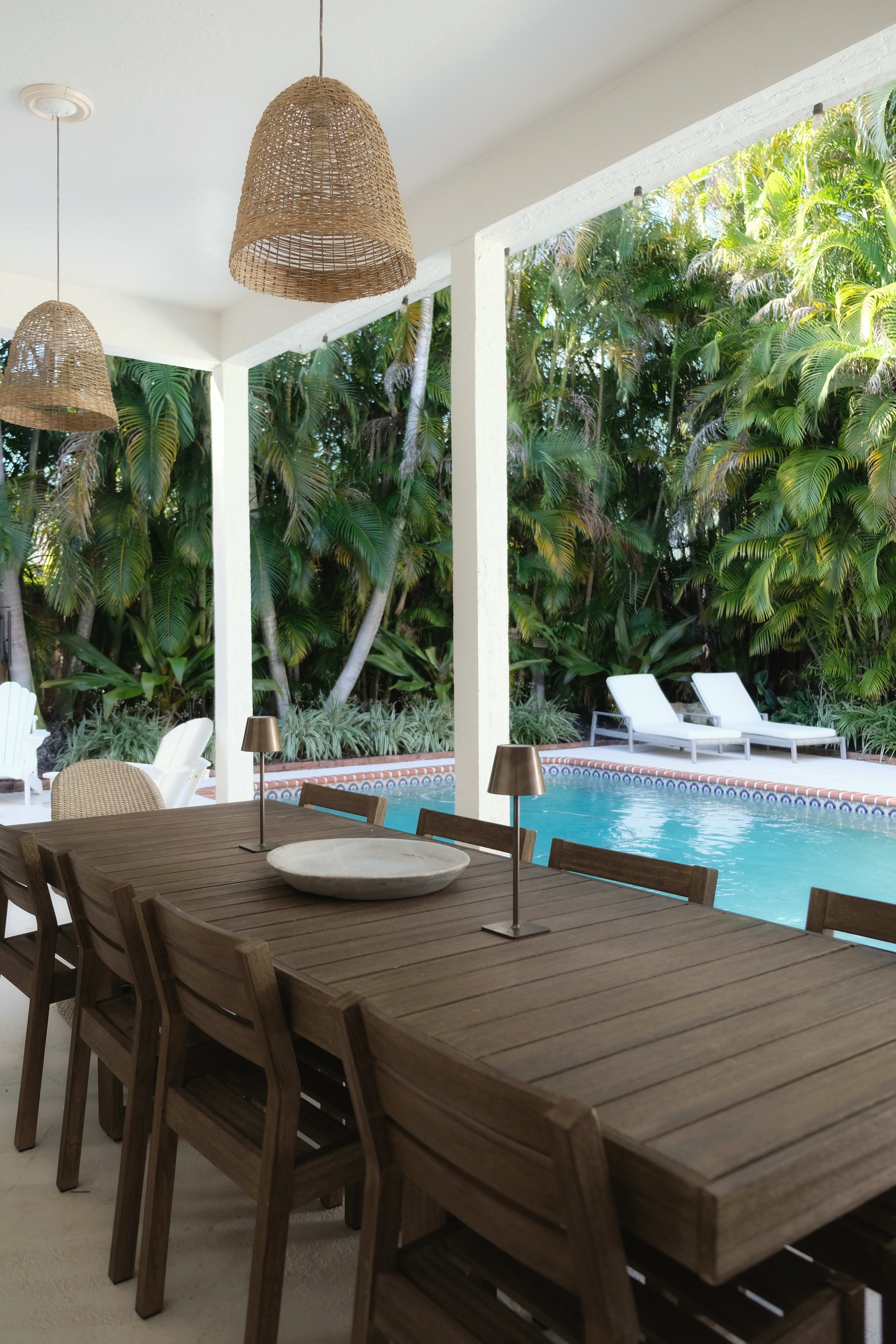 A wooden dining table with chairs is set under hanging woven lamps beside a swimming pool surrounded by tropical plants.