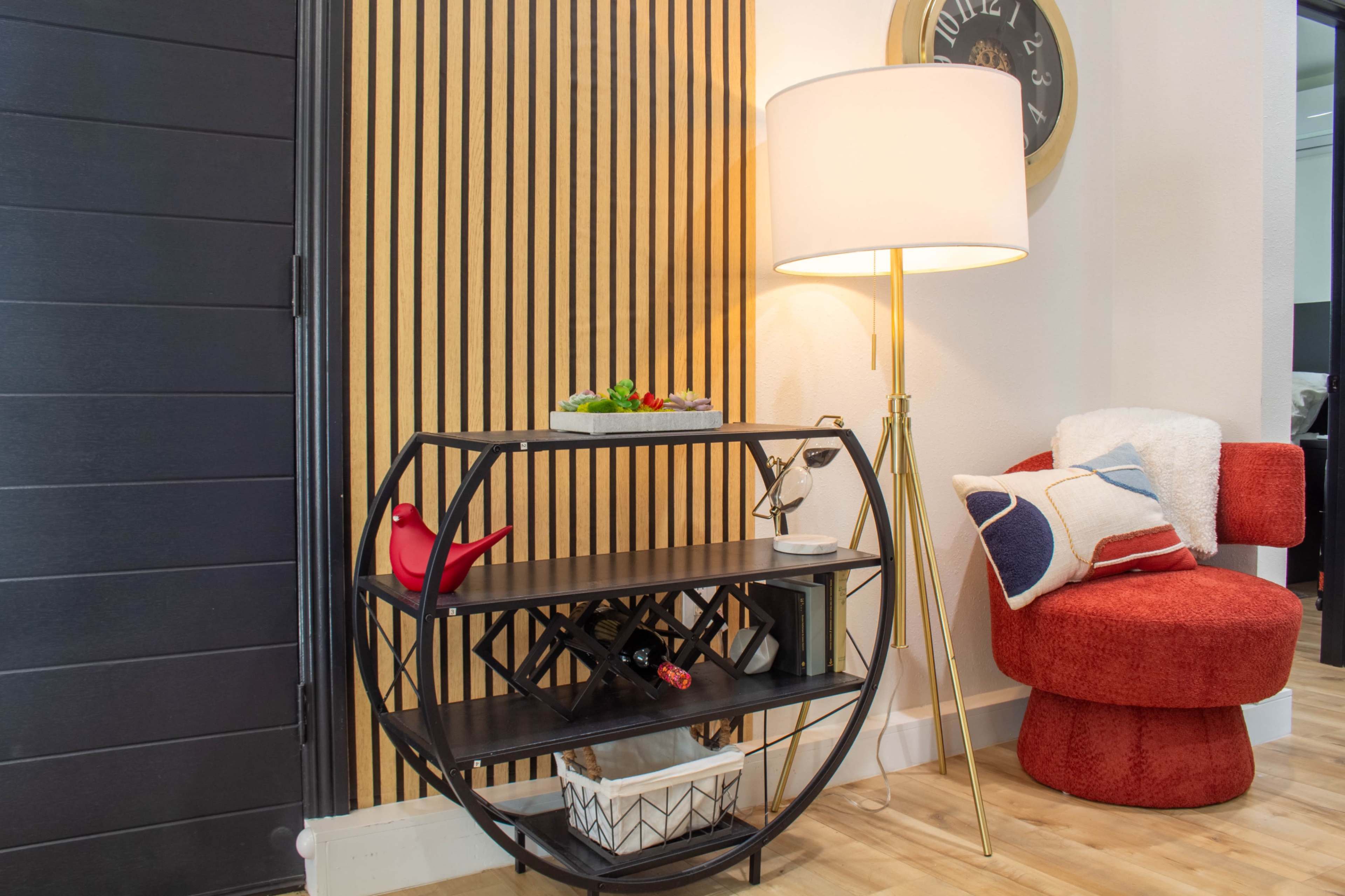 A modern living area features a black metal shelving unit with a decorative display, a floor lamp, and a red upholstered chair against a striped wood wall.