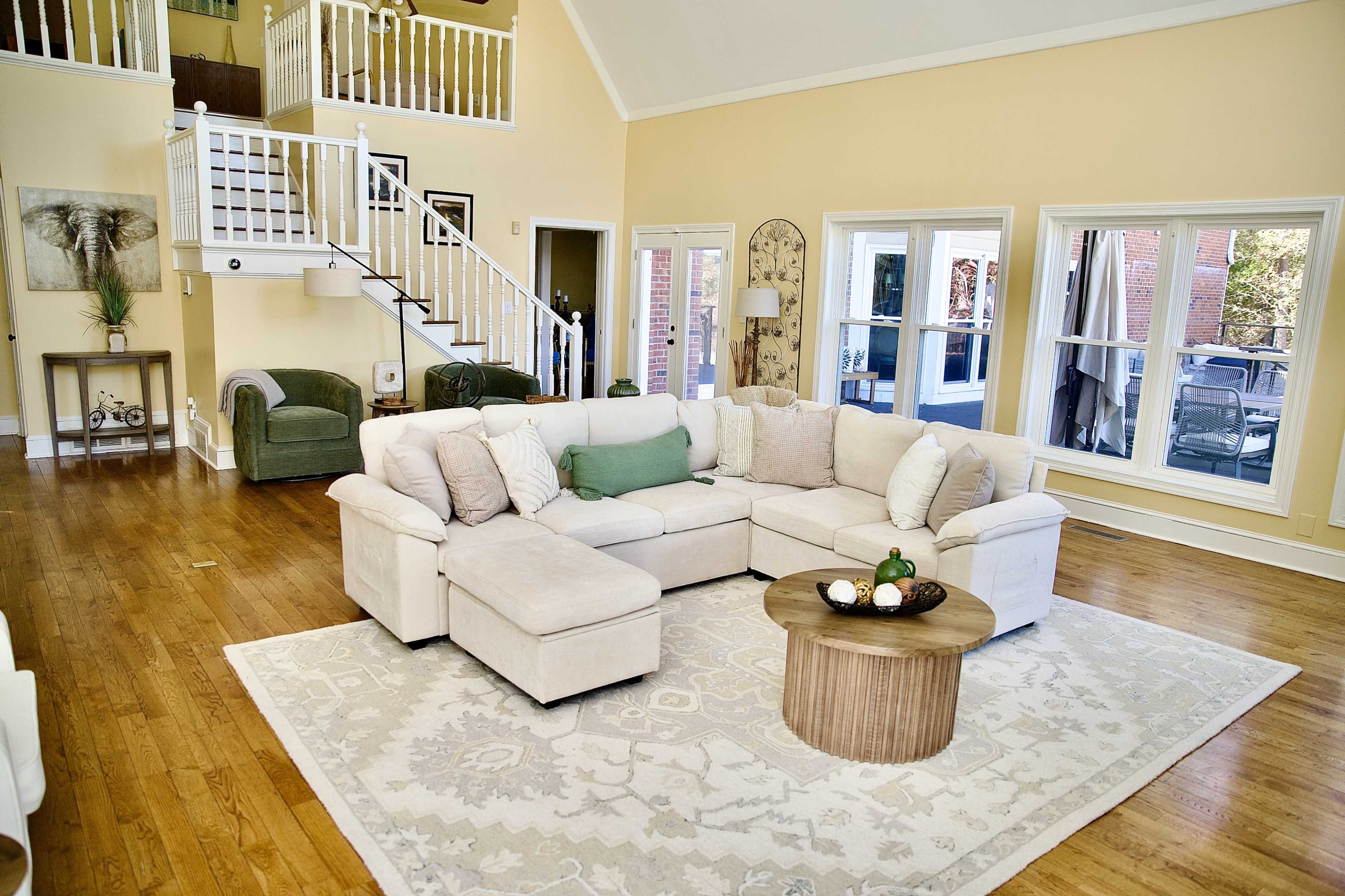 The image shows a spacious, well-lit living room featuring a sectional sofa, a wooden coffee table, a large area rug, and a staircase leading to an upper level.