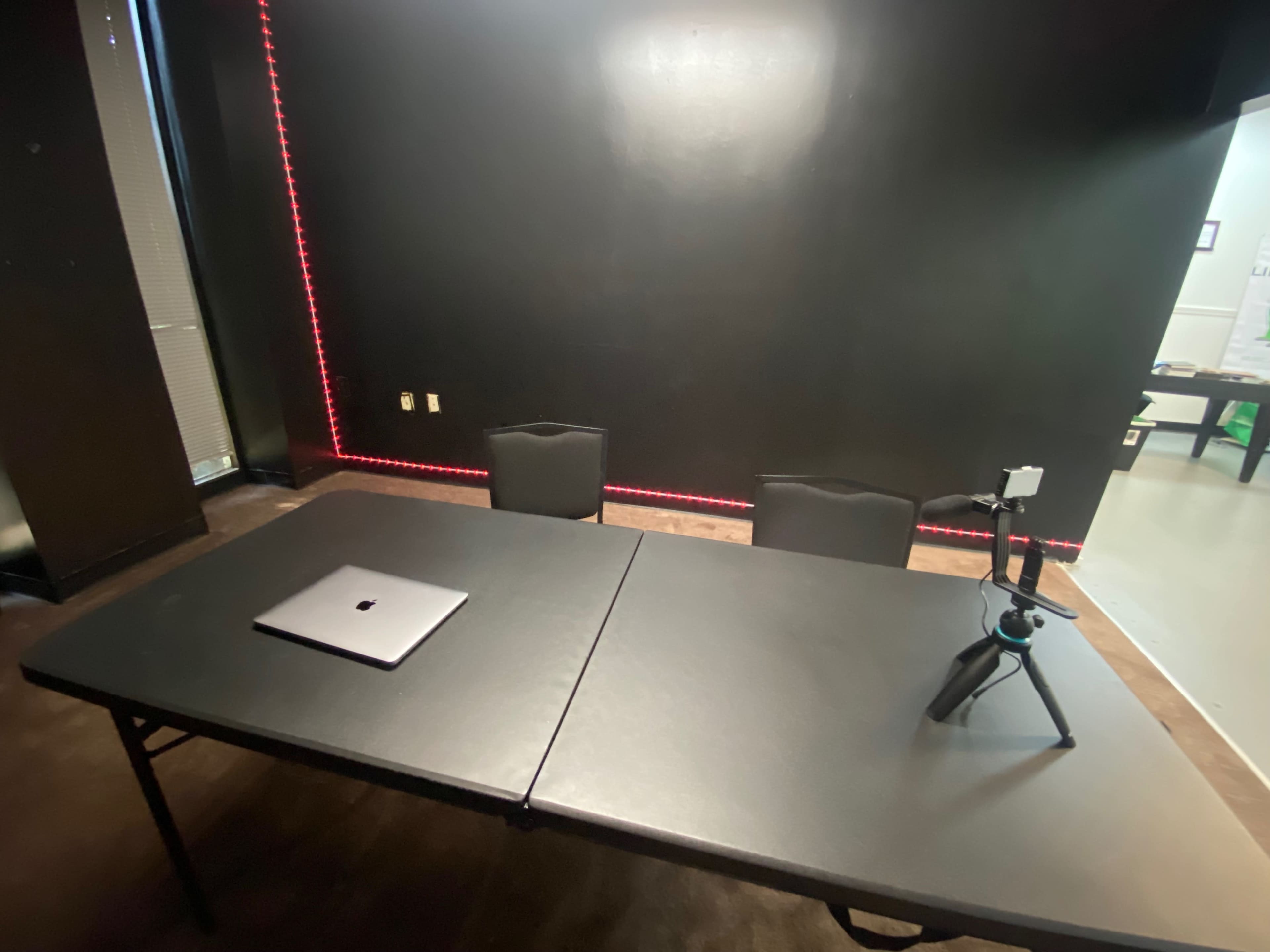 The image shows a sparse room with a black wall, a table at the center, two black chairs, a laptop on the table, and a tripod next to it.