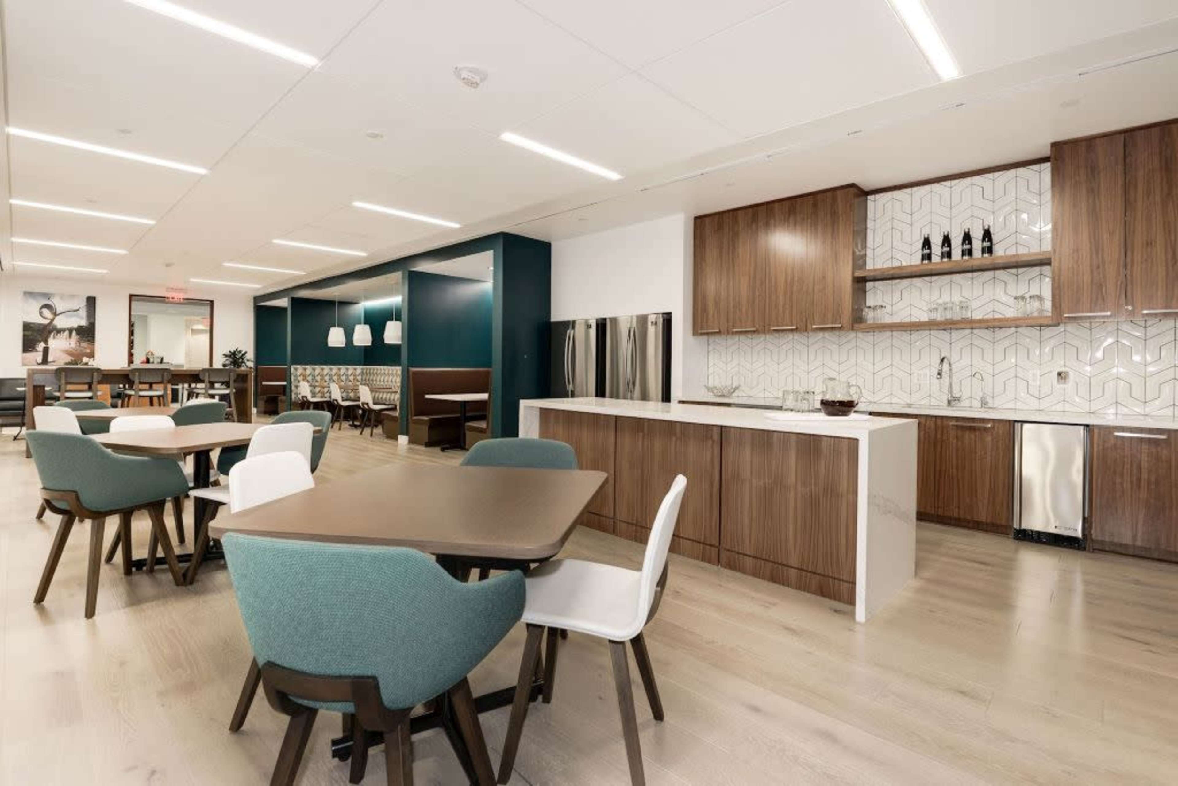 A modern kitchen and dining area features wooden cabinetry, white countertops, and a dining space with a mix of chairs around several tables.