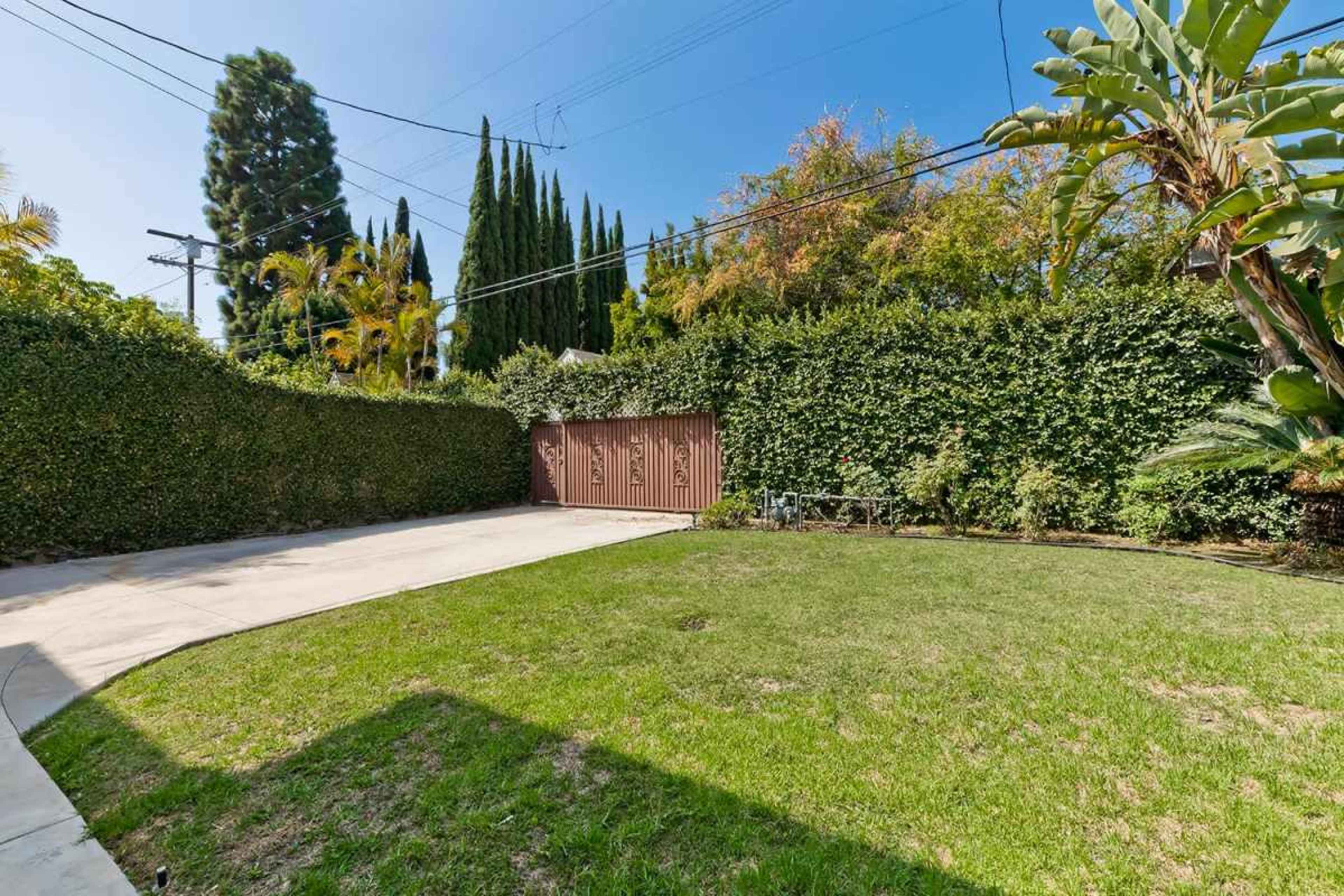 The image shows a spacious backyard with green grass, surrounded by tall hedges and trees, and featuring a brown gate.