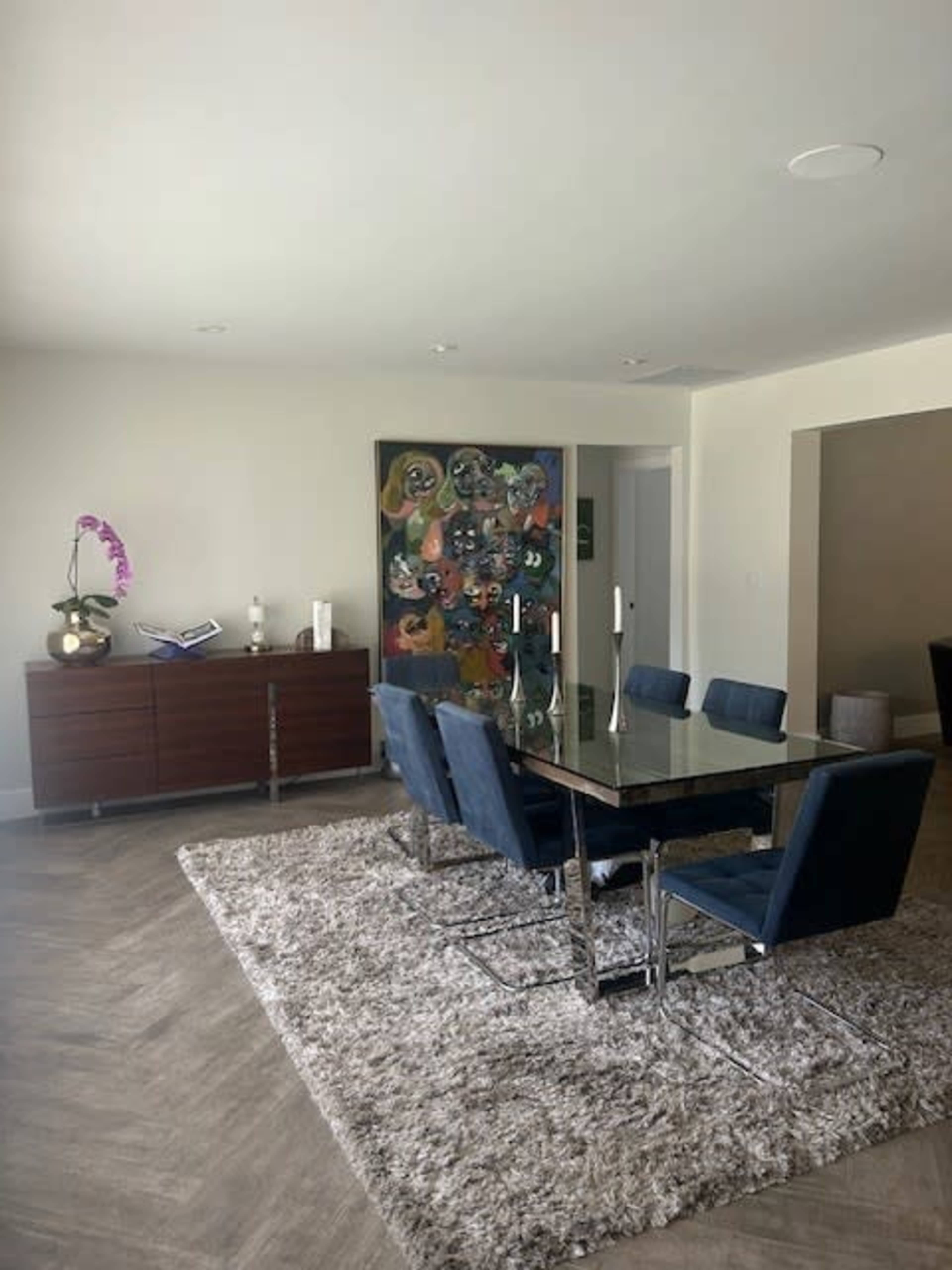 A modern dining area features a glass table surrounded by blue upholstered chairs, with a wooden sideboard and colorful artwork on the wall.