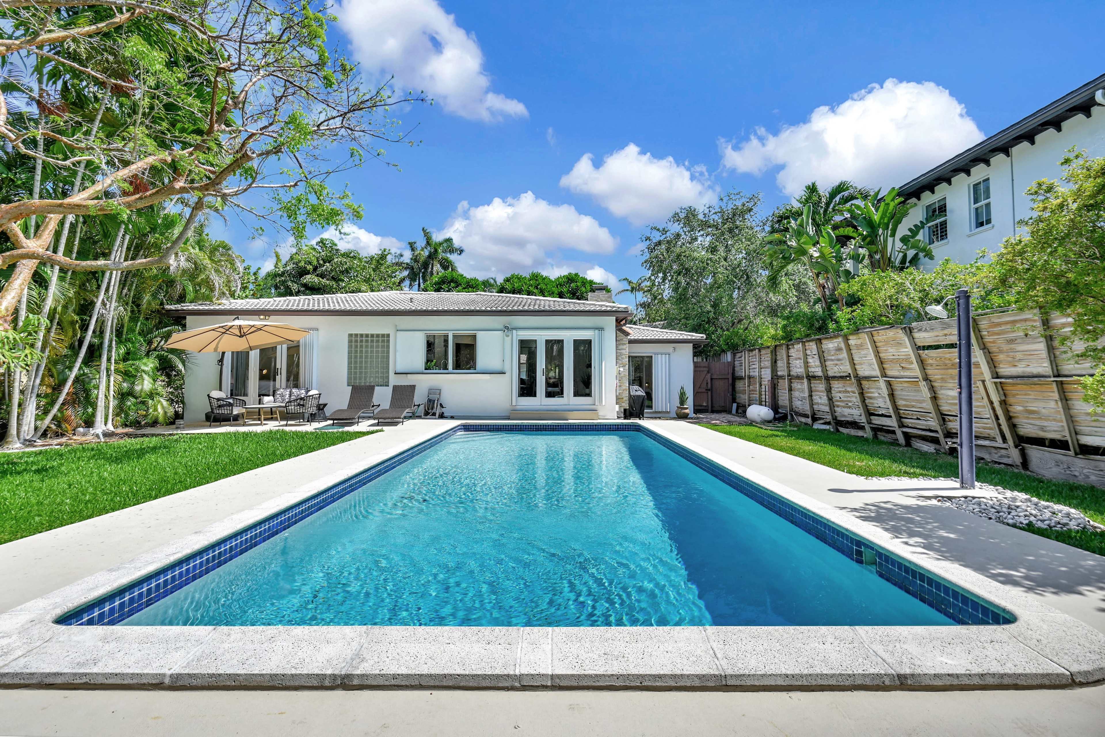 A modern house with a swimming pool and lounge area is surrounded by greenery and palm trees.