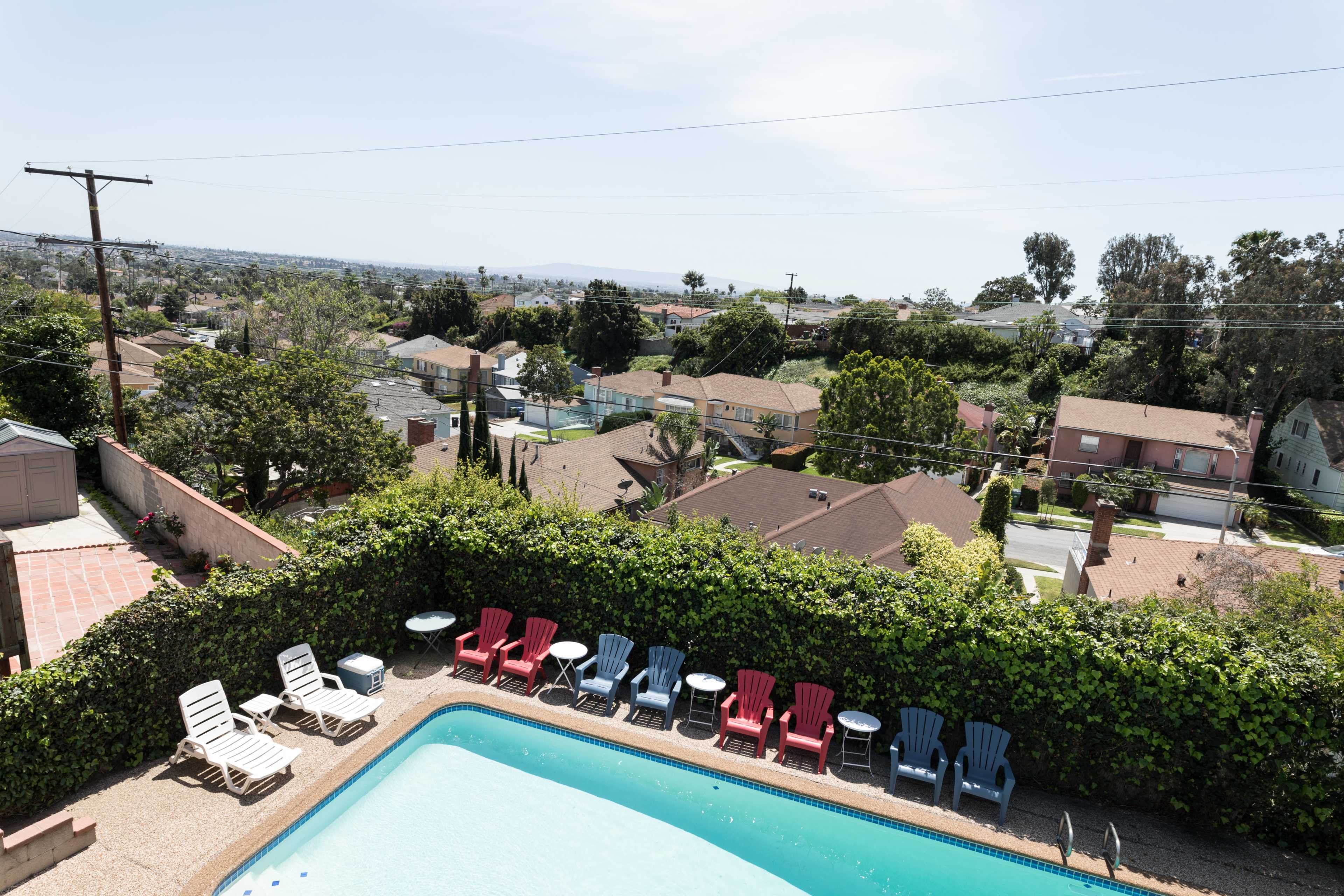 The image shows a swimming pool surrounded by colorful lounge chairs, with a view of houses and trees in the background under a clear sky.