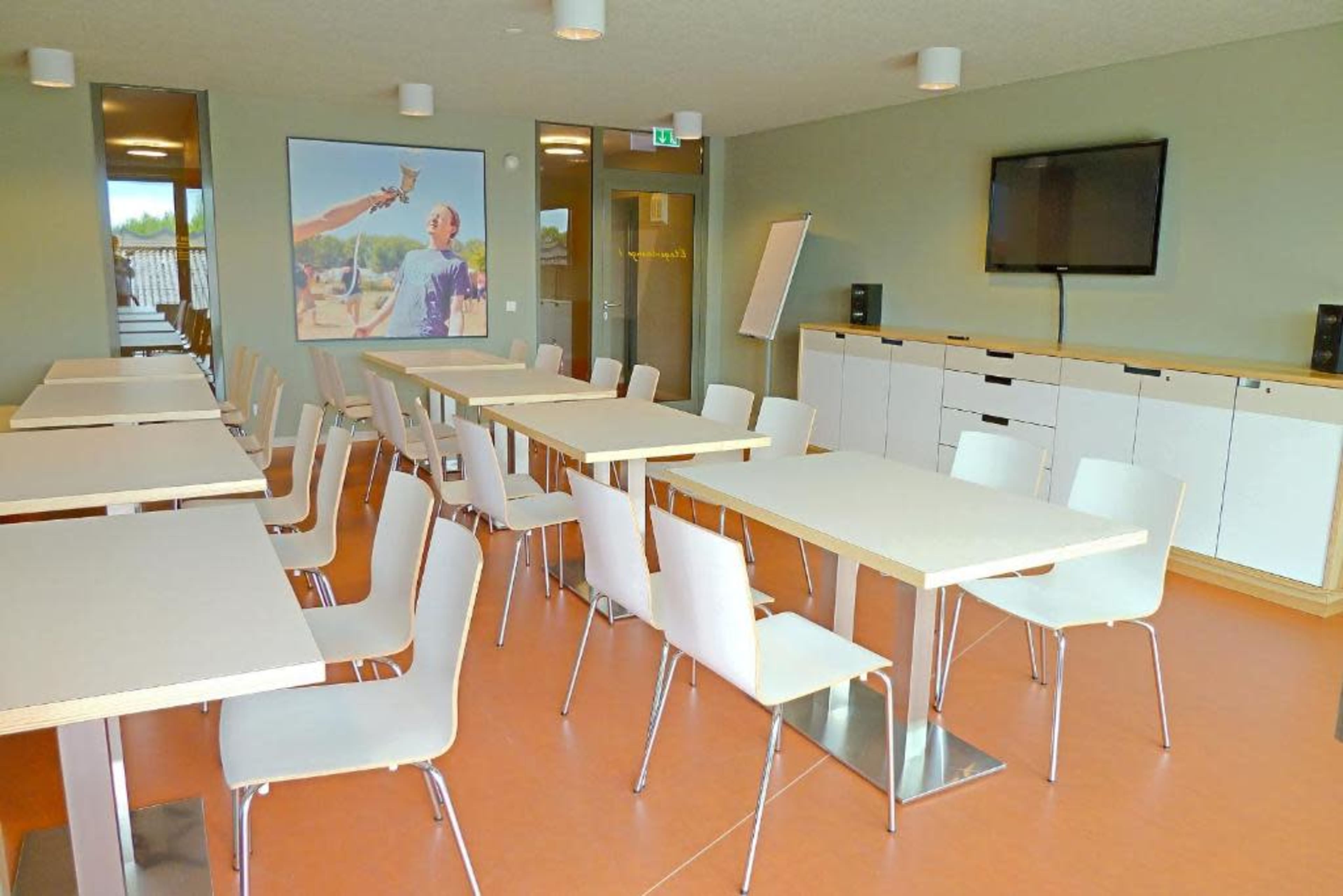 A modern conference room with several tables and chairs arranged neatly, and a large TV screen mounted on the wall.