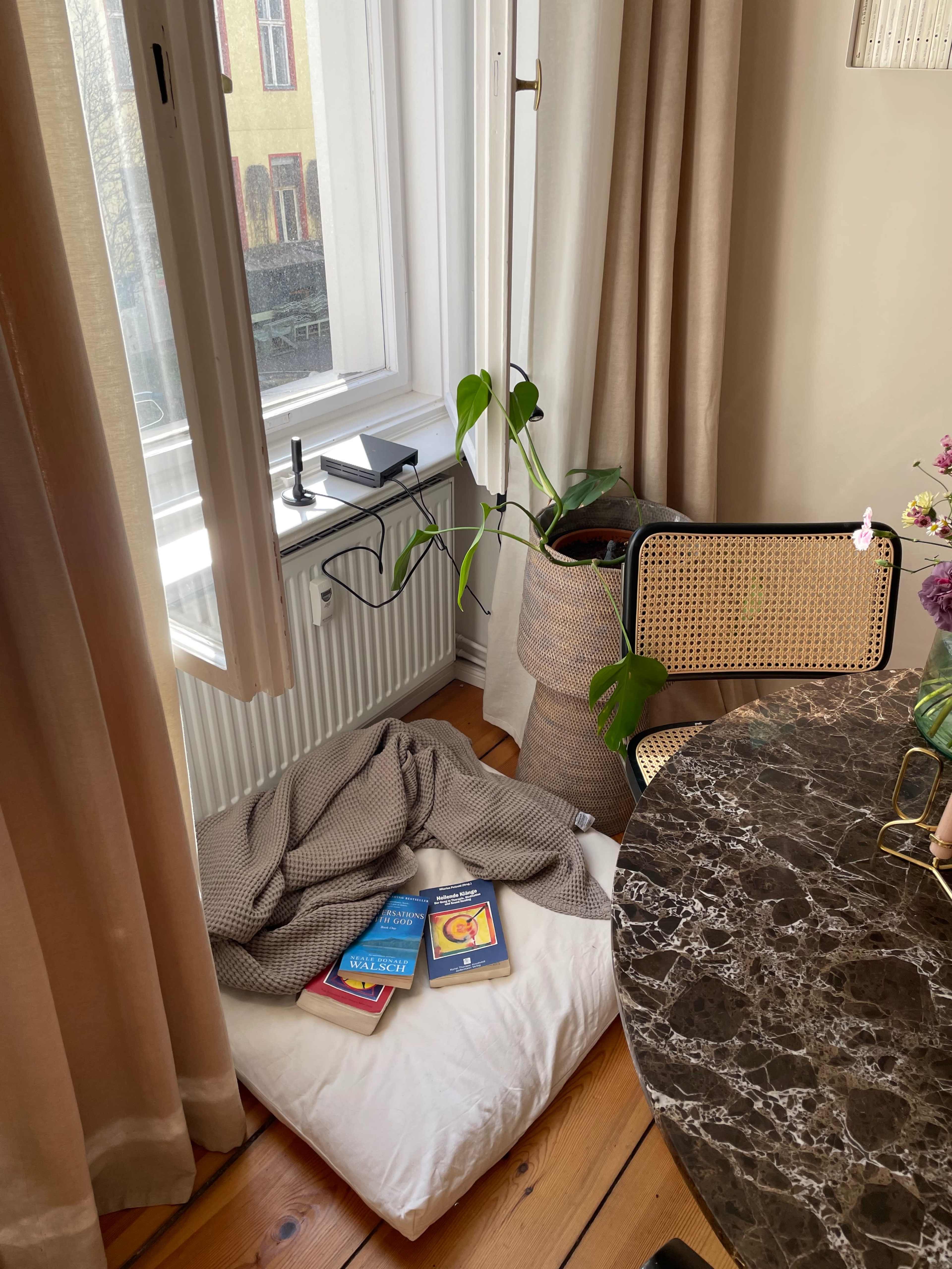 A white cushion on the wooden floor beside a potted plant and a marble table, with several books stacked on top.
