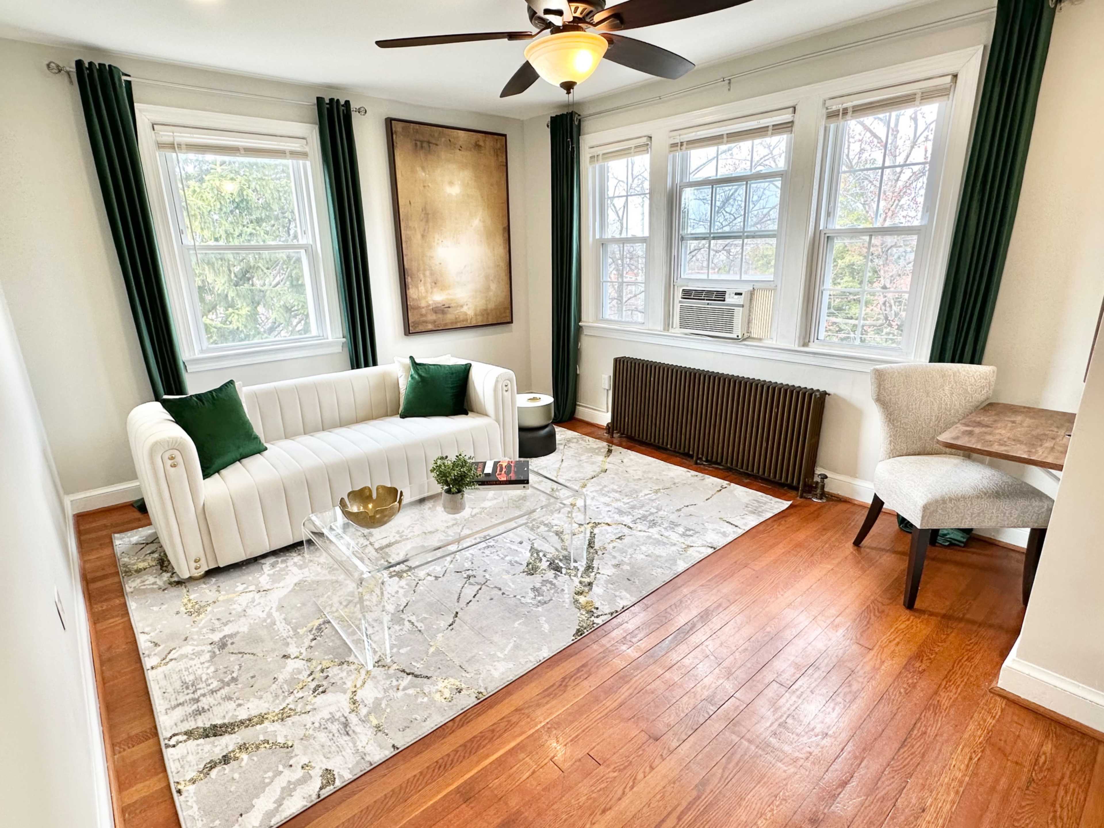 The room features a white sofa with green cushions, a transparent glass coffee table, and a light gray area rug, set against a backdrop of large windows and hardwood flooring.
