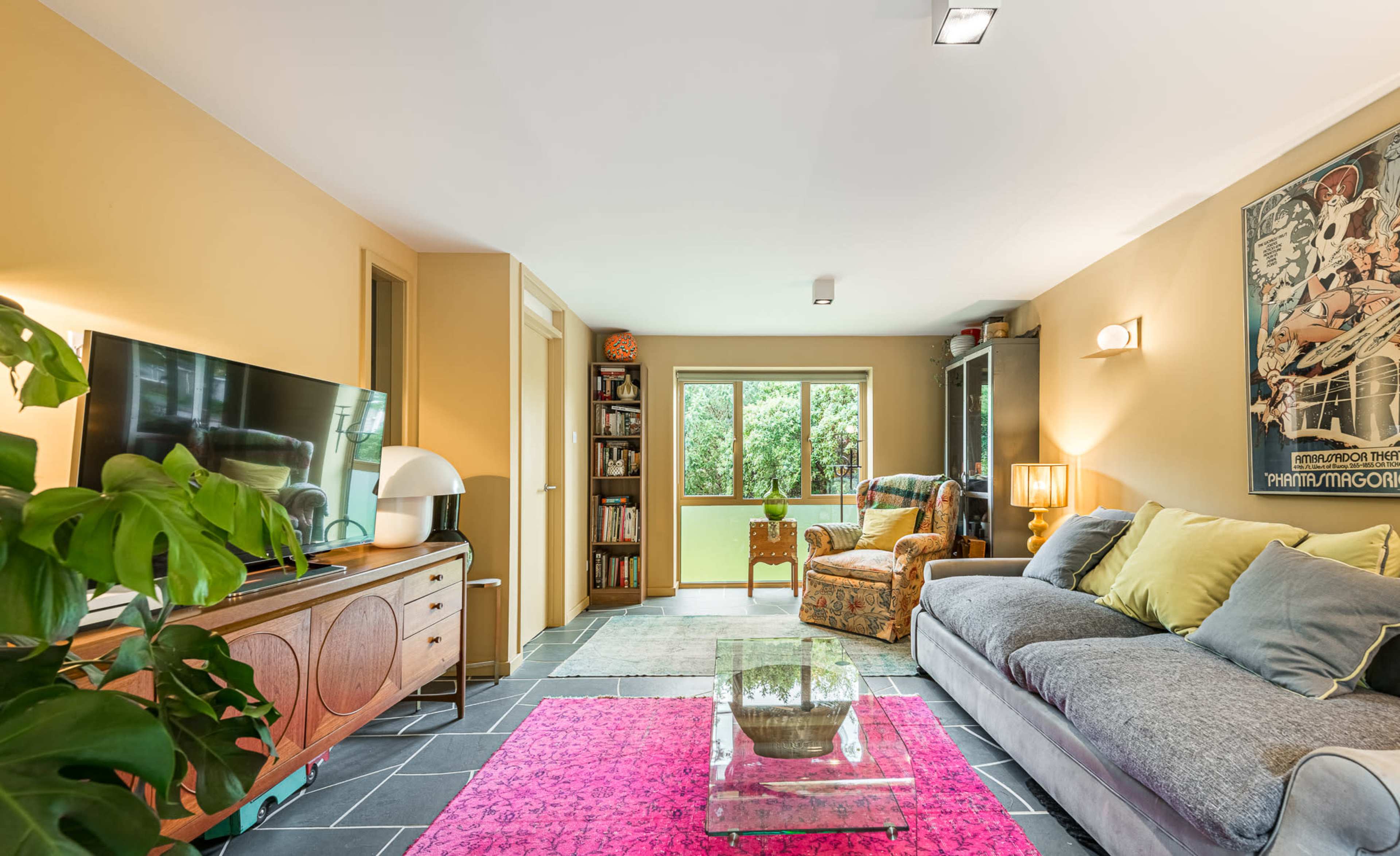 The image shows a cozy living room with a gray sofa, a colorful area rug, a wooden TV stand, and large windows that allow natural light to enter.