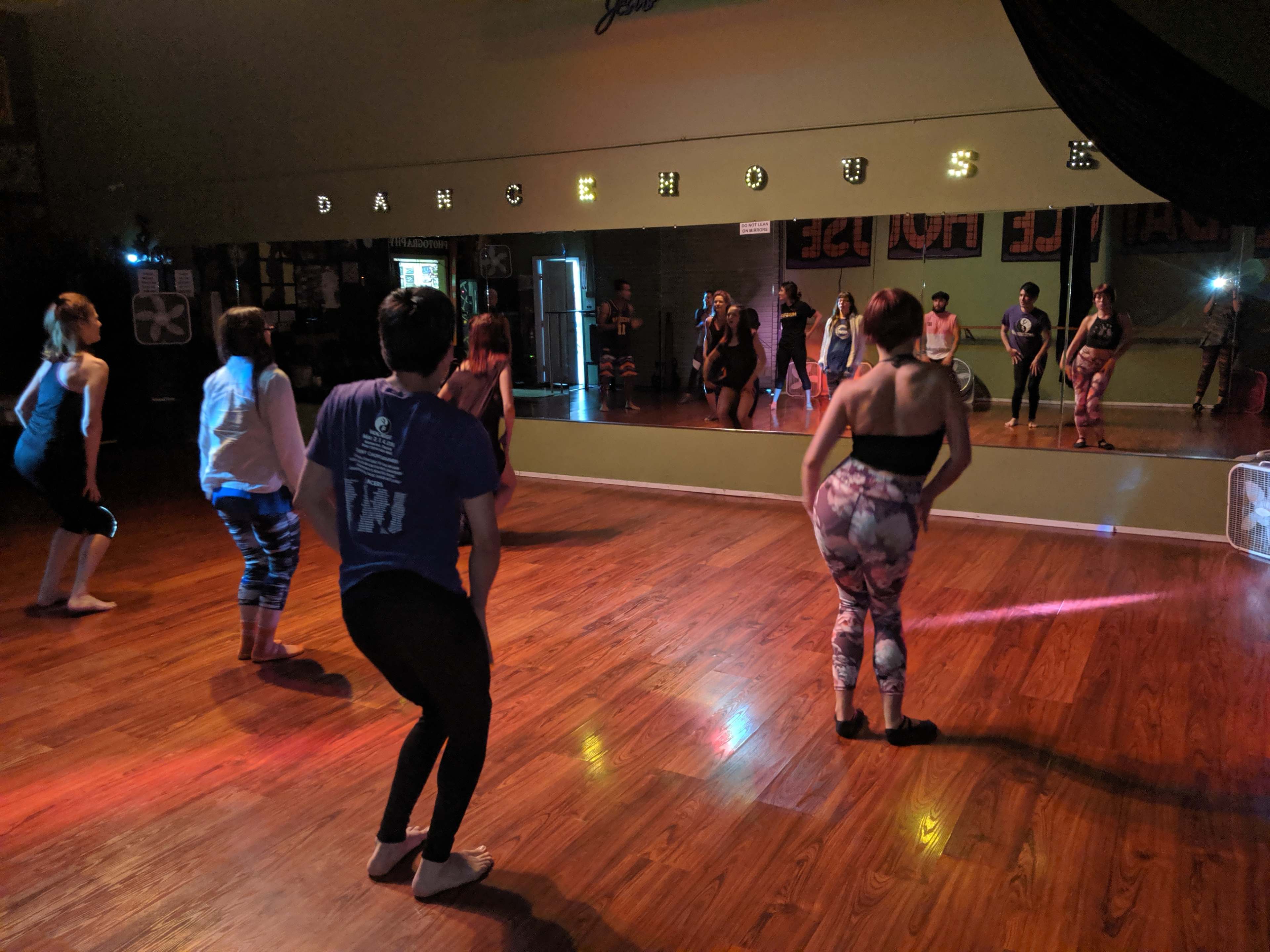 A group of dancers practices in a studio with wooden floors and a large mirror.