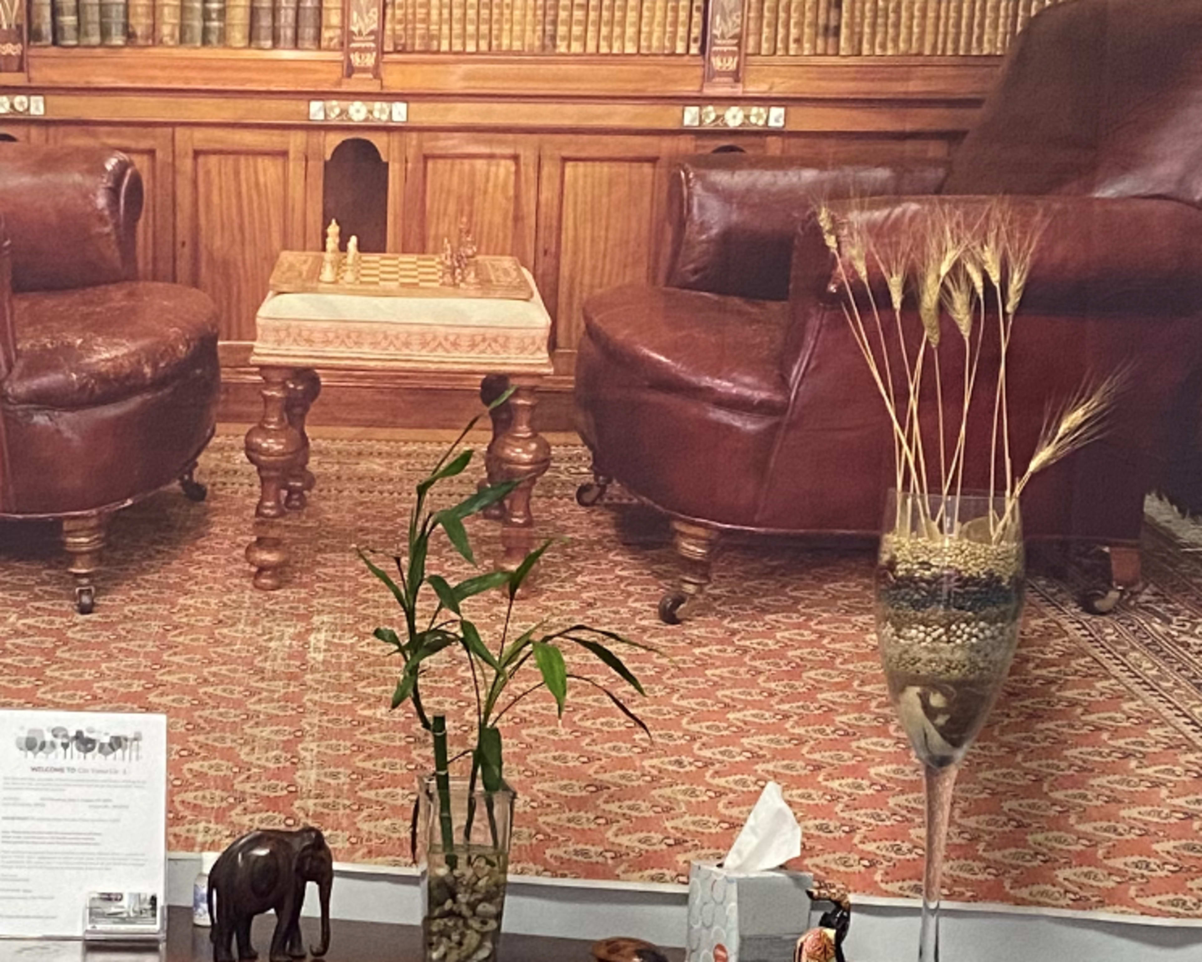 A cozy room with two leather chairs facing a chessboard, set against a backdrop of wooden bookshelves and a patterned rug.