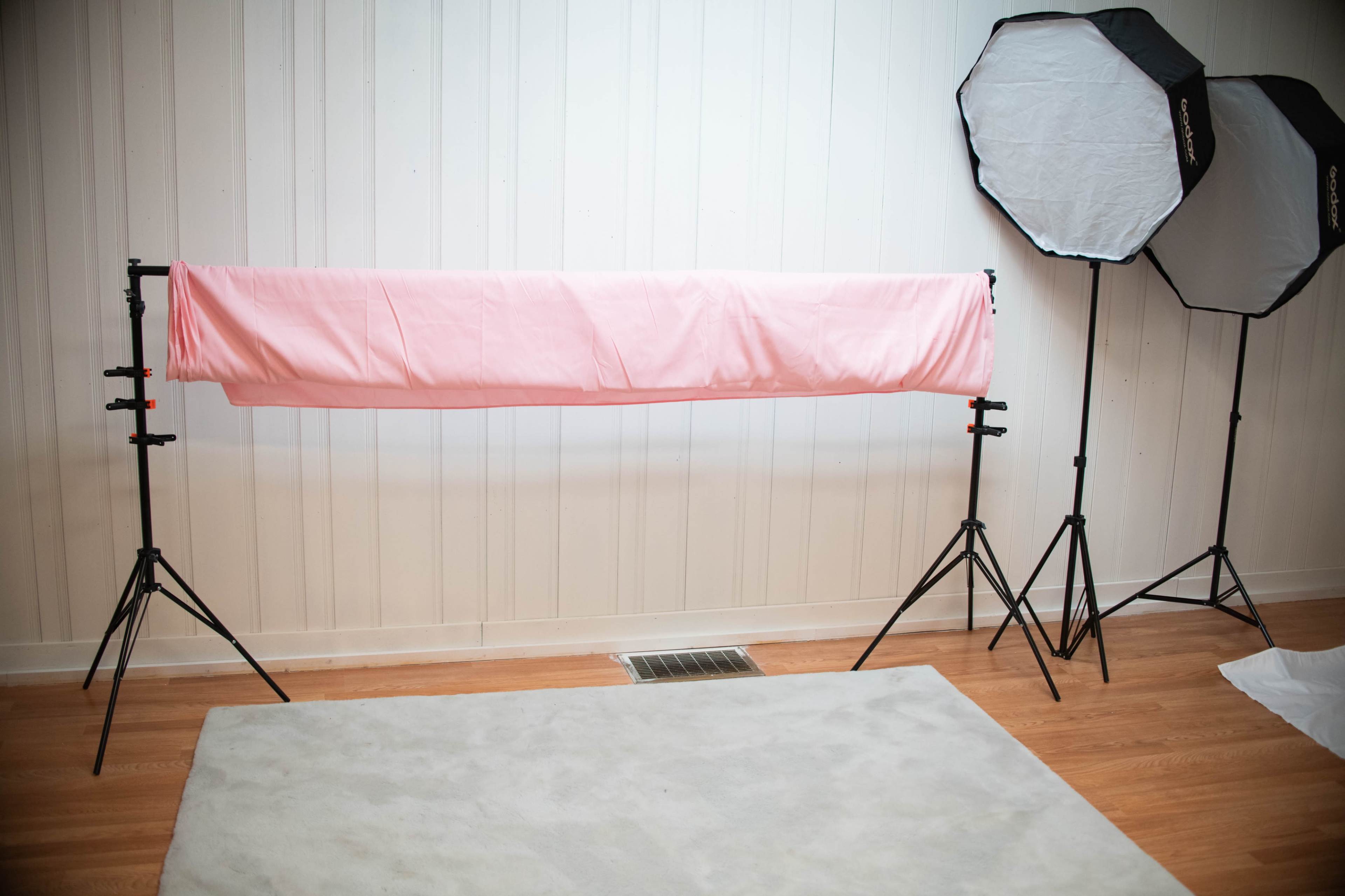A photography setup features a pink backdrop stretched between two stands, flanked by two softbox lights, on a wooden floor.