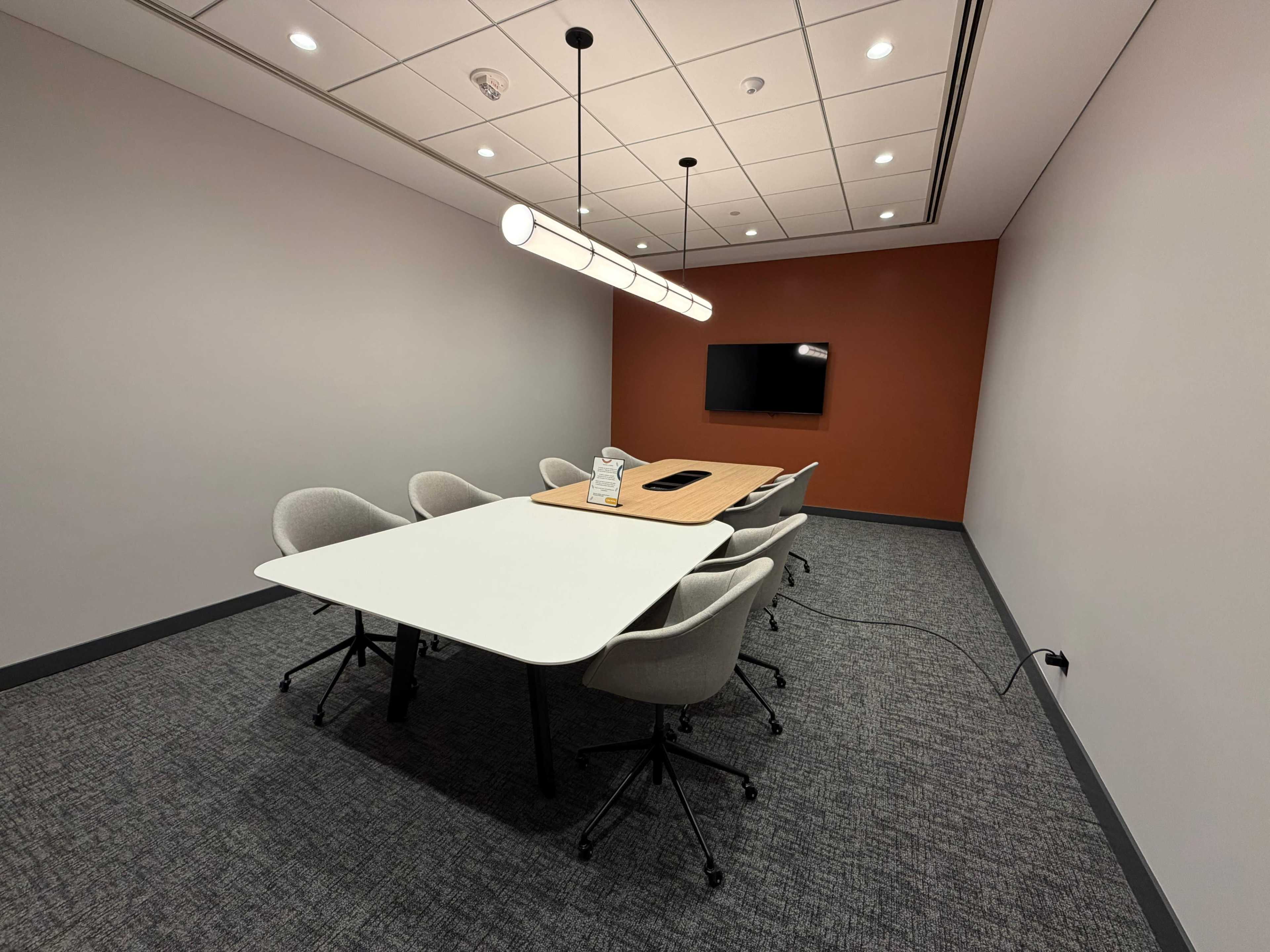 A conference room features a rectangular table surrounded by six chairs, with a mounted screen on a dark orange wall and overhead lighting.