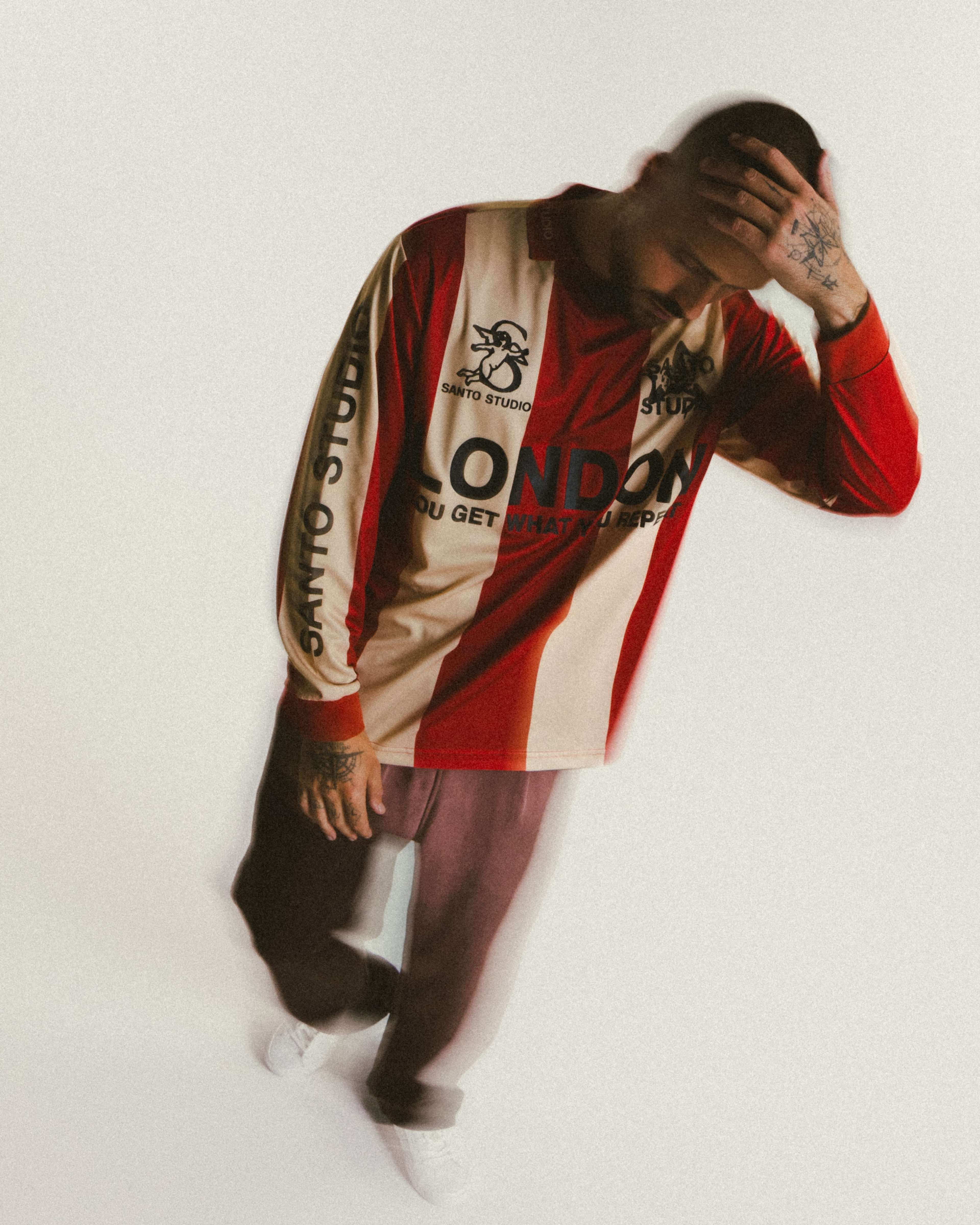 A person wearing a long-sleeve striped shirt with the word "LONDON" prominently displayed stands with their head bowed, appearing contemplative.