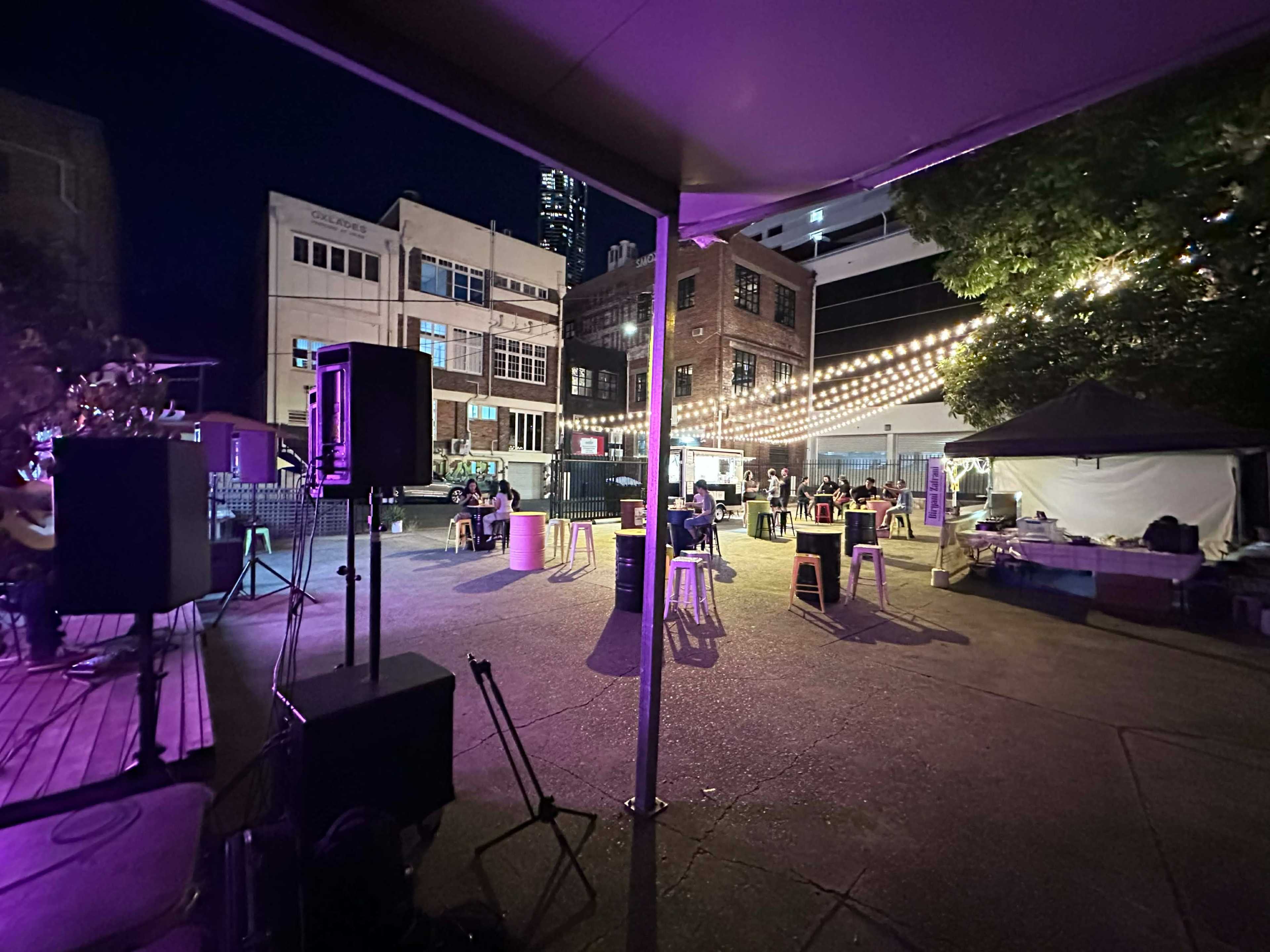 The image shows an outdoor evening scene with string lights illuminating a gathering space, where people are seated at tables and a small stage setup is visible.