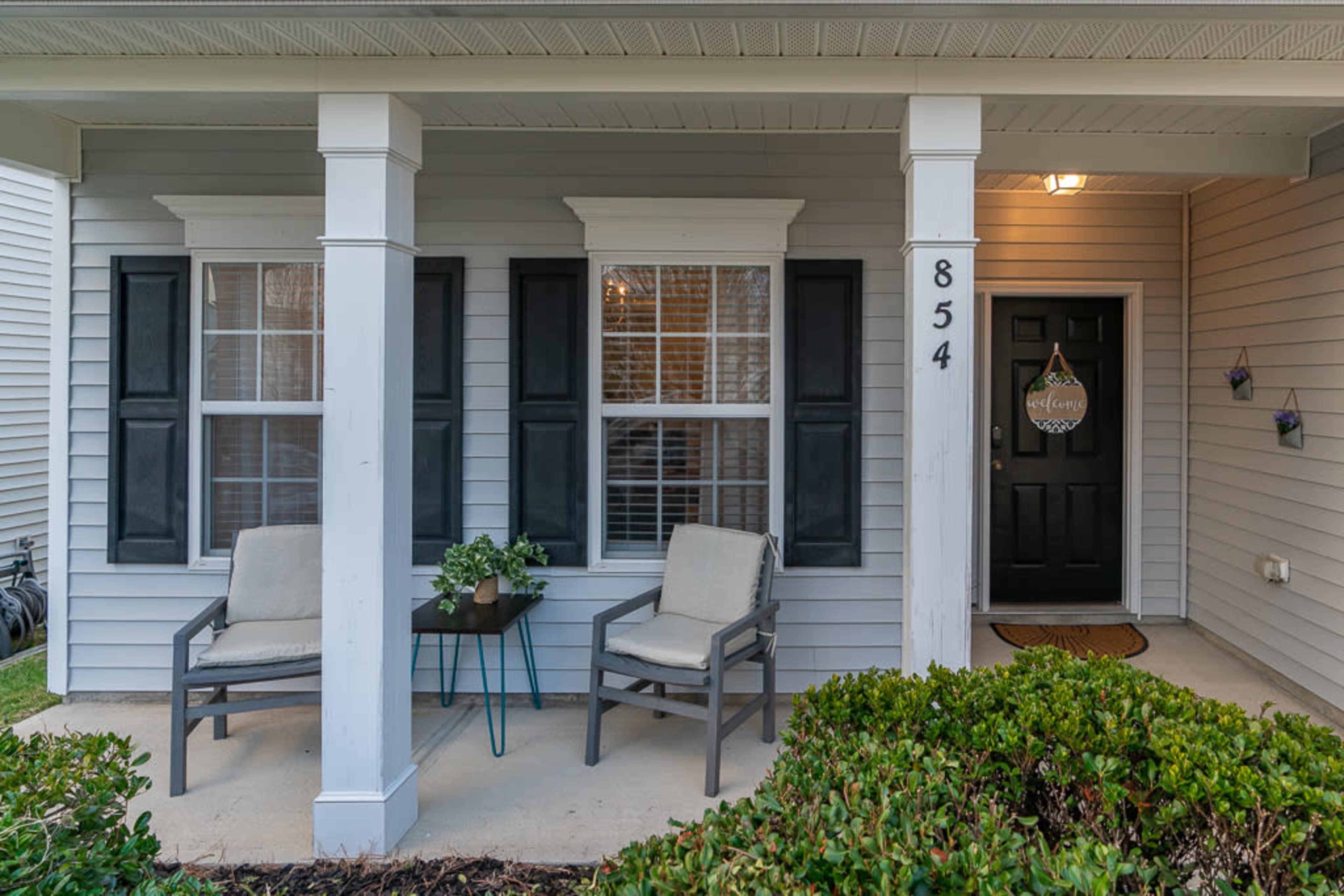A front porch with two gray chairs, a small table, and welcoming decor on the door, framed by two white pillars.