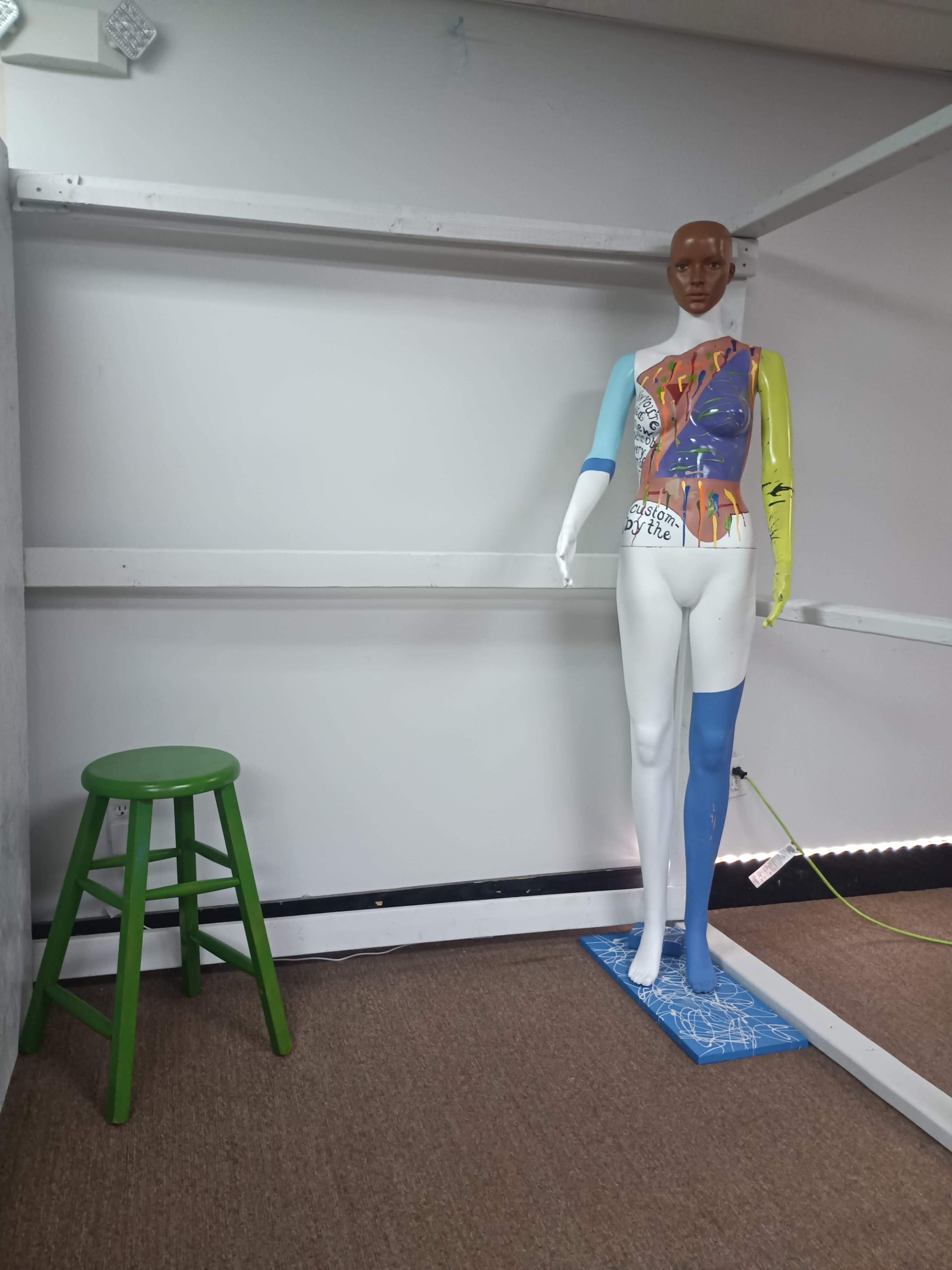 A painted mannequin stands beside a green wooden stool against a bare white wall.