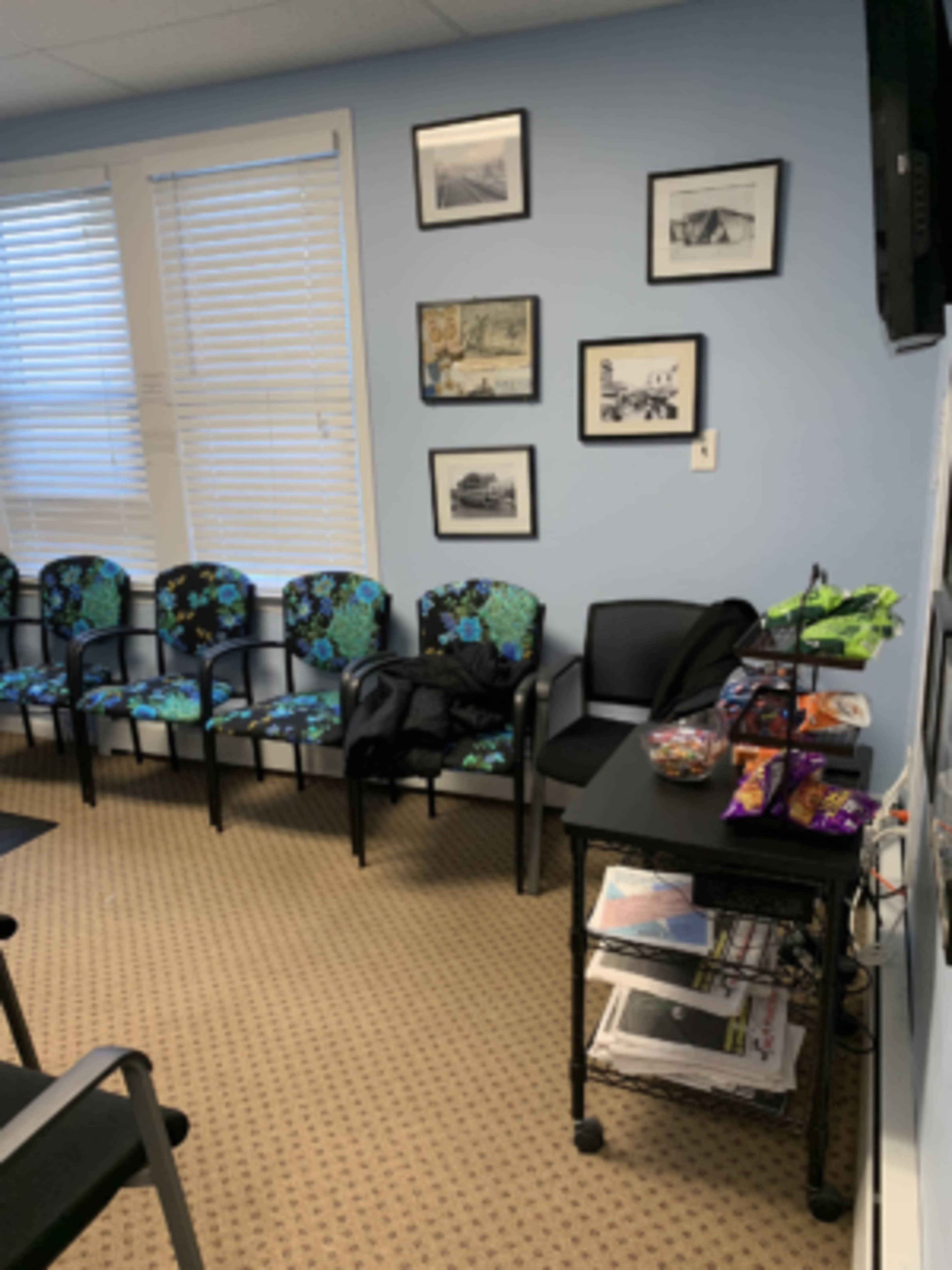 The image shows a waiting room with several patterned chairs, a table covered with snacks and magazines, and framed pictures on the wall.