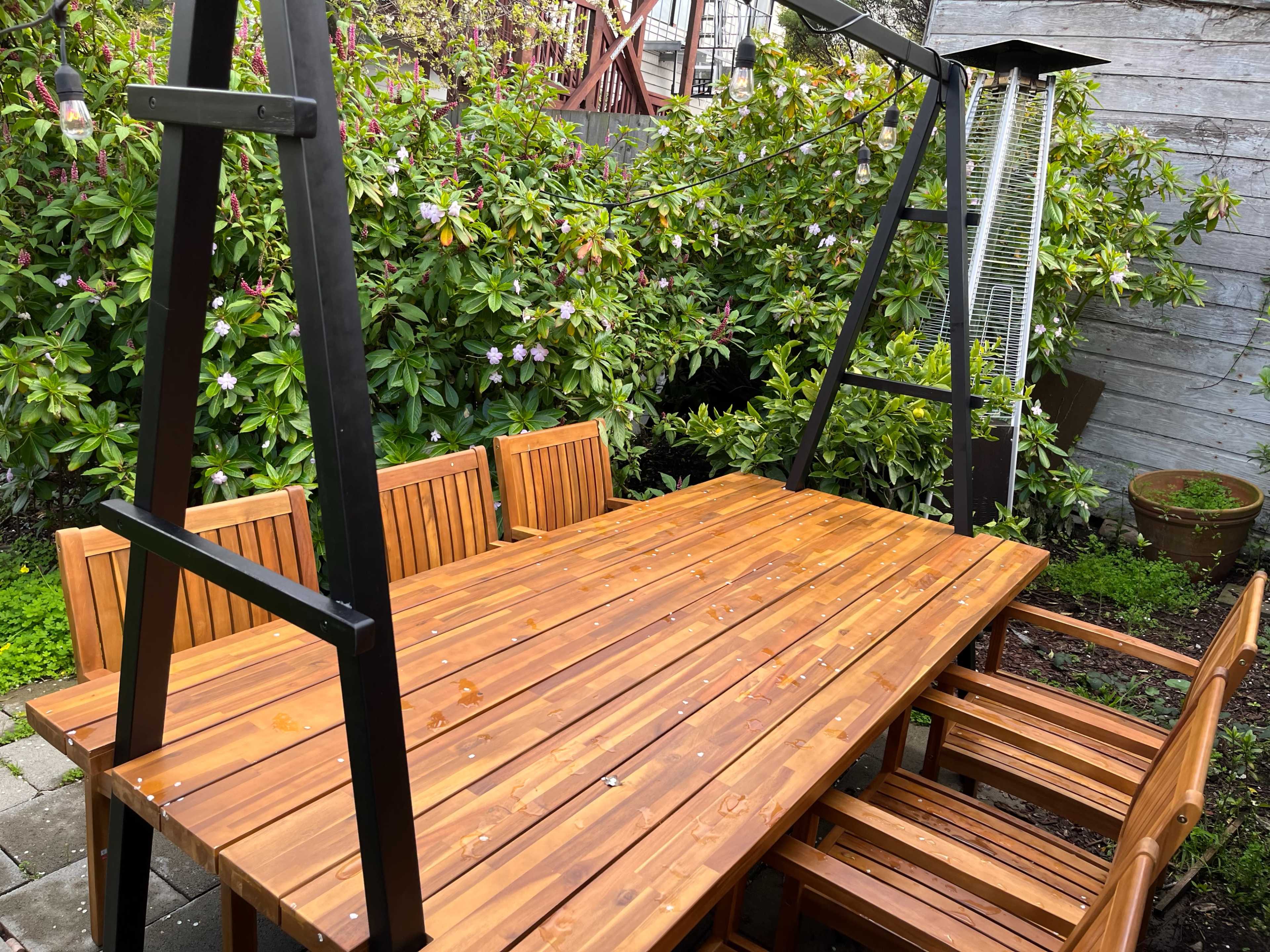 A wooden outdoor dining table with matching chairs is set up in a garden surrounded by flowering shrubs.