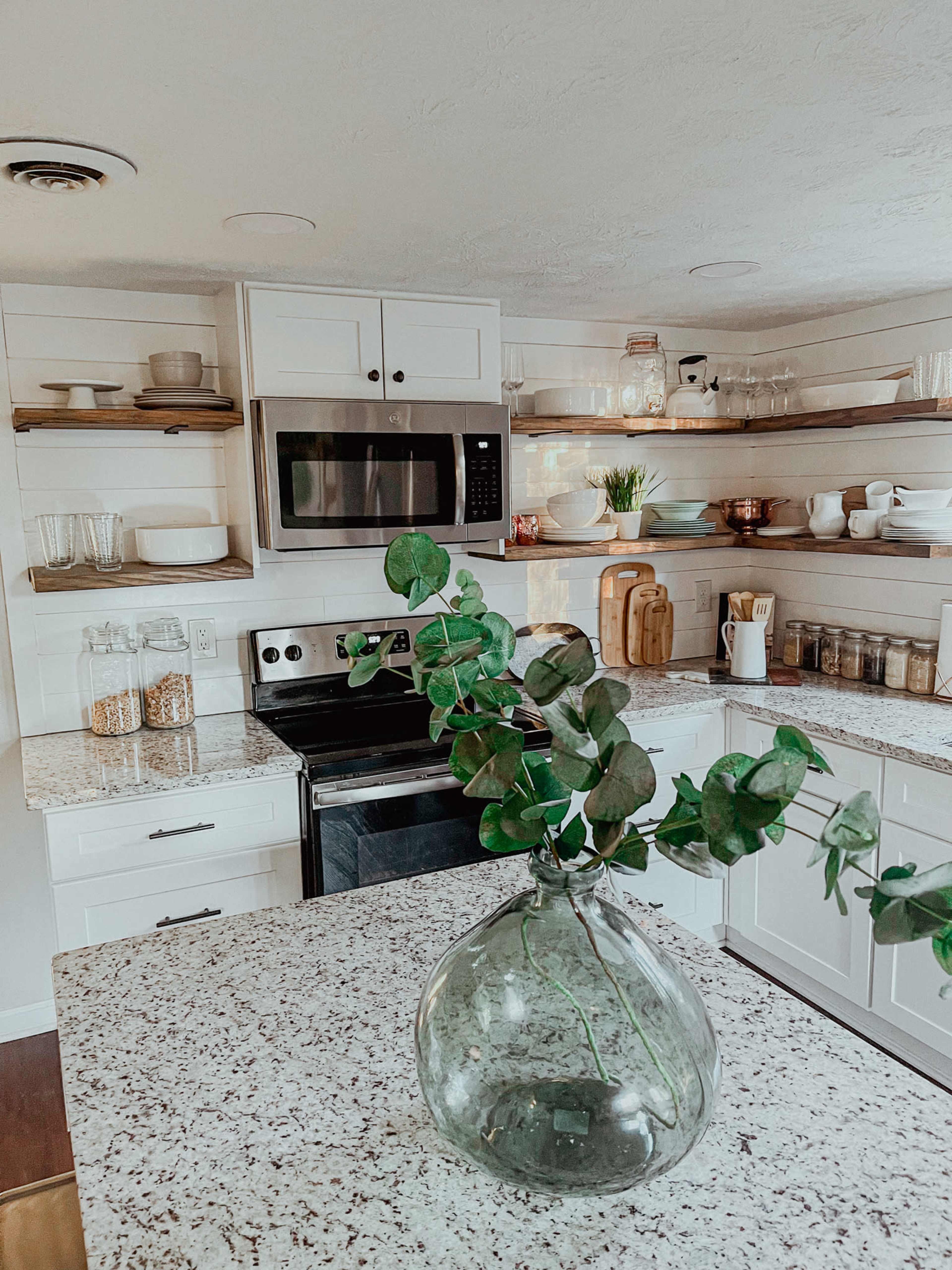 A modern kitchen features white cabinets, open shelving with various dishes and glassware, and a stone countertop with a decorative glass vase and greenery.
