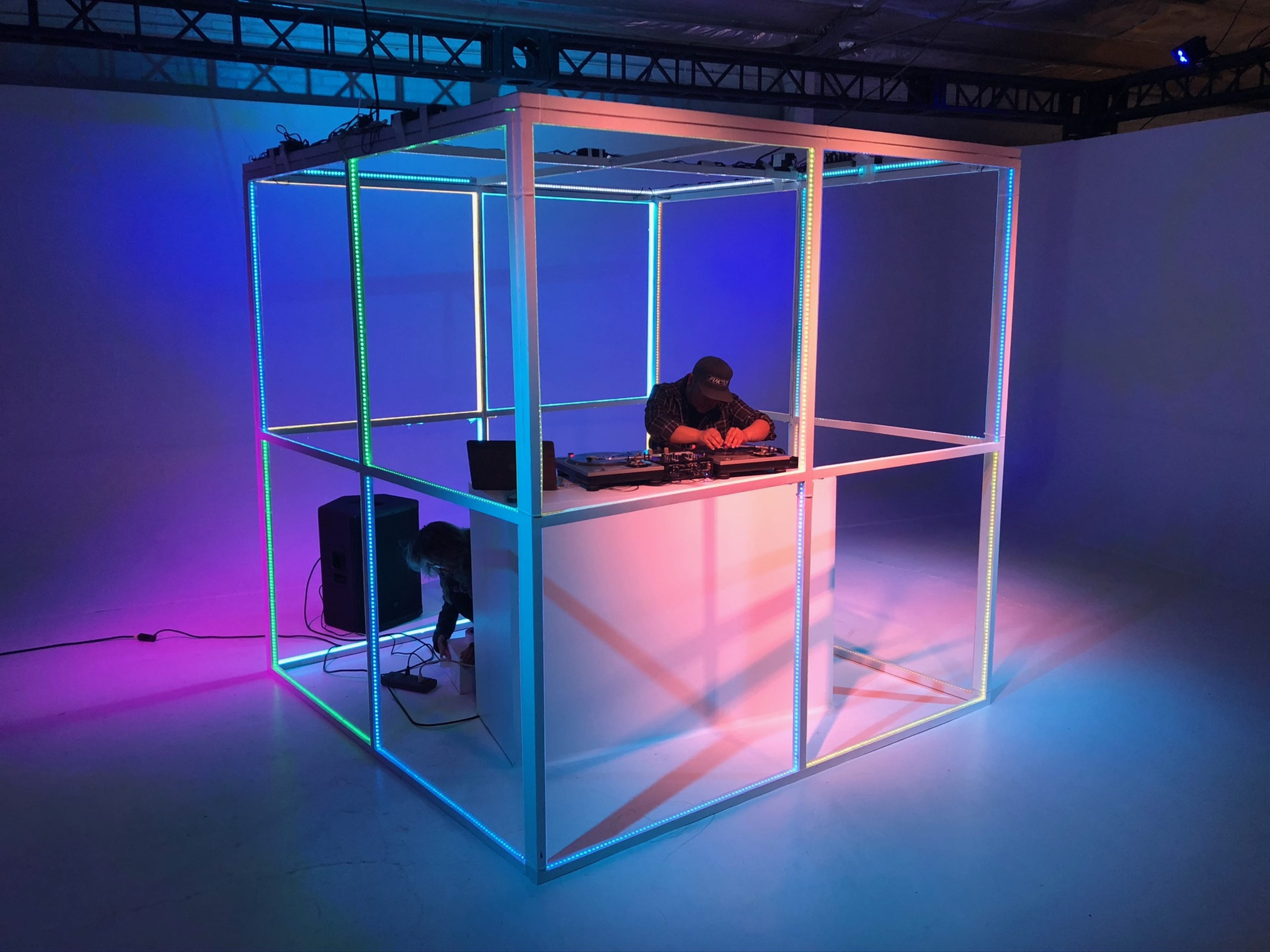 A DJ performs inside a lighted, clear structure with colorful illumination in a spacious, uncluttered setting.