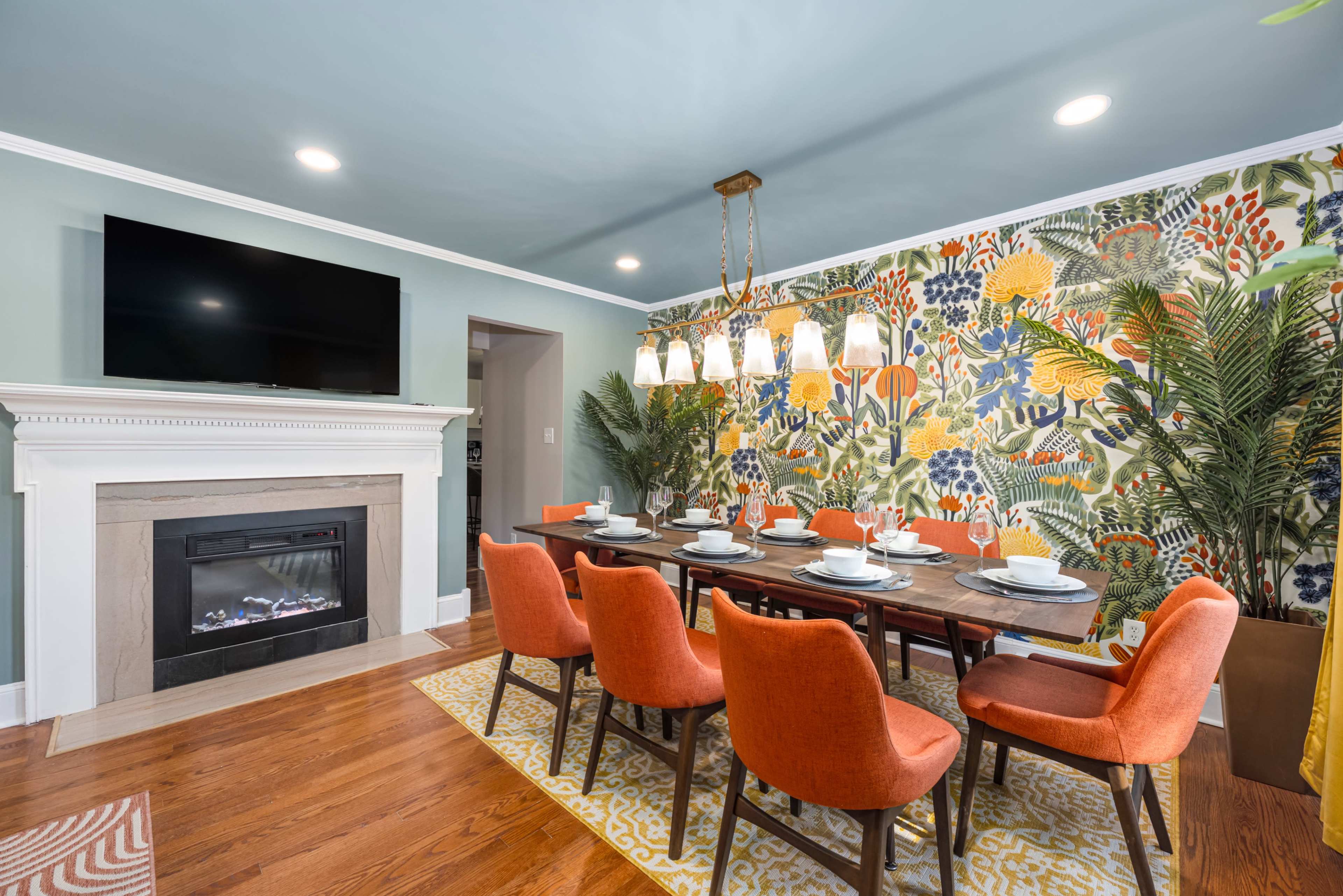 The image shows a bright dining room featuring a wooden table set with dishes, surrounded by orange chairs, against a floral-patterned accent wall and a fireplace with a television mounted above it.
