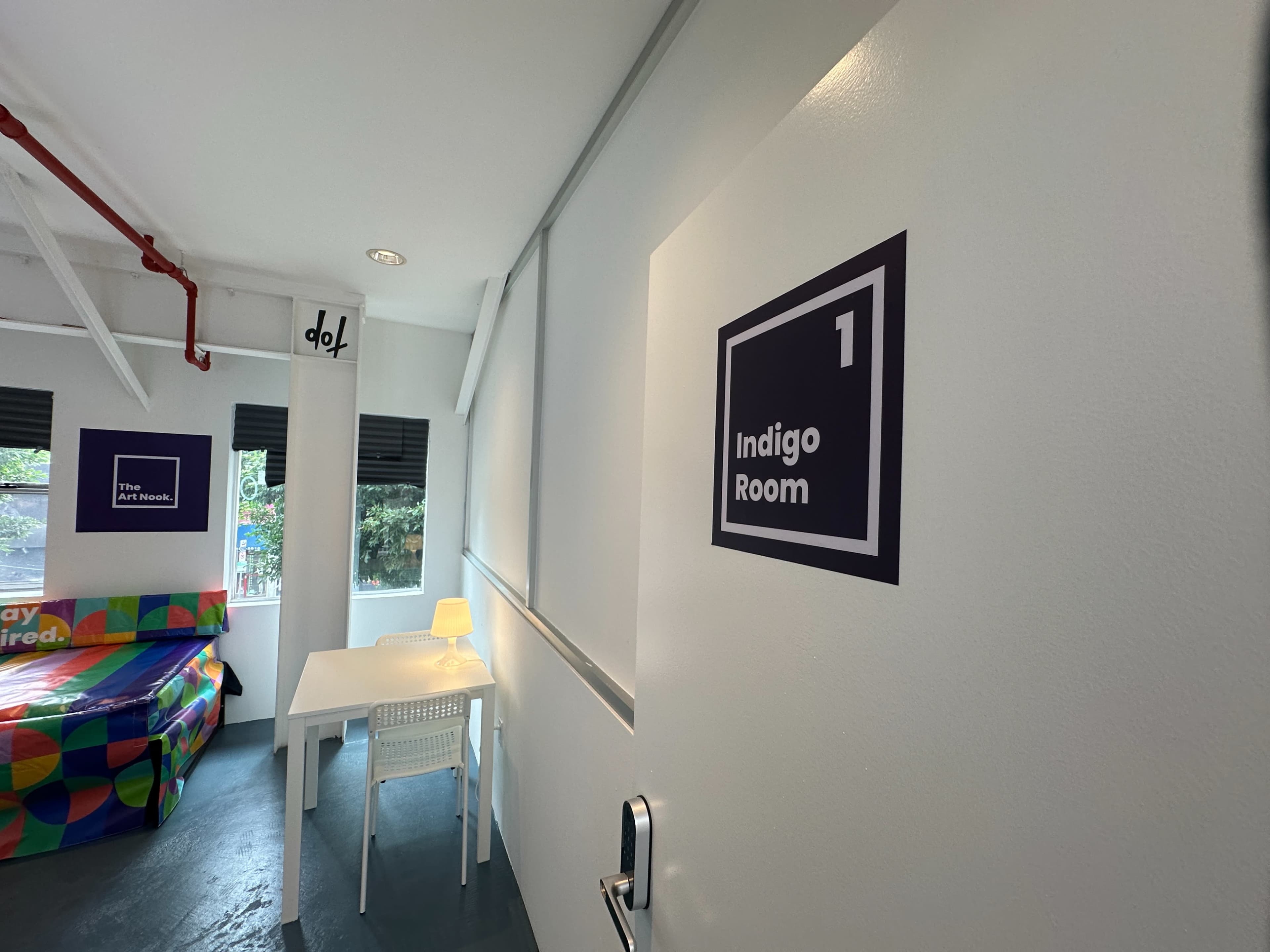 The image shows a room labeled "Indigo Room" with a sign on the wall, a small table and chair, and a colorful couch in the background.