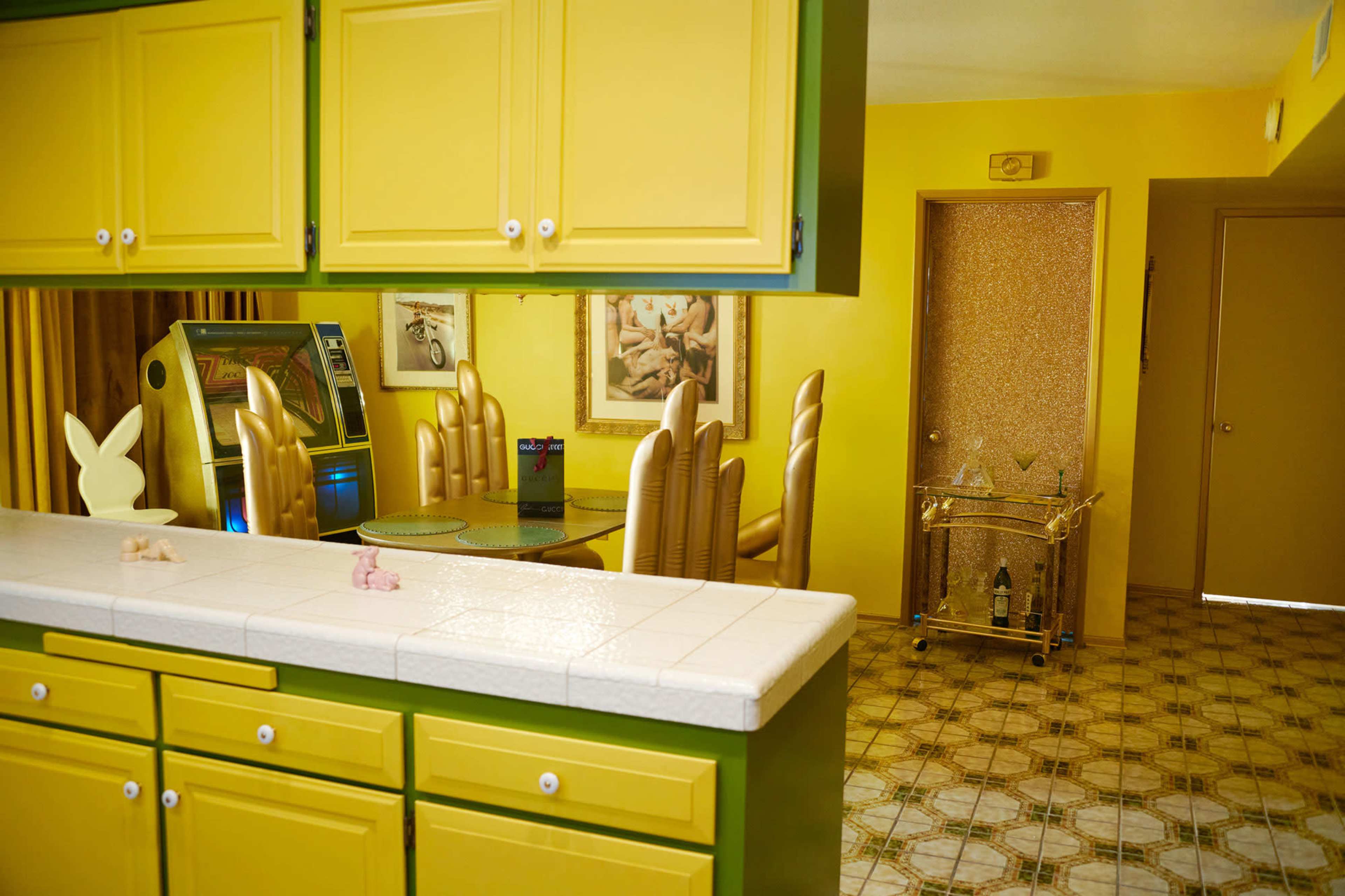 A brightly colored kitchen with yellow cabinets and a tiled floor, featuring a dining area with gold-colored chairs and an adjacent bar cart.