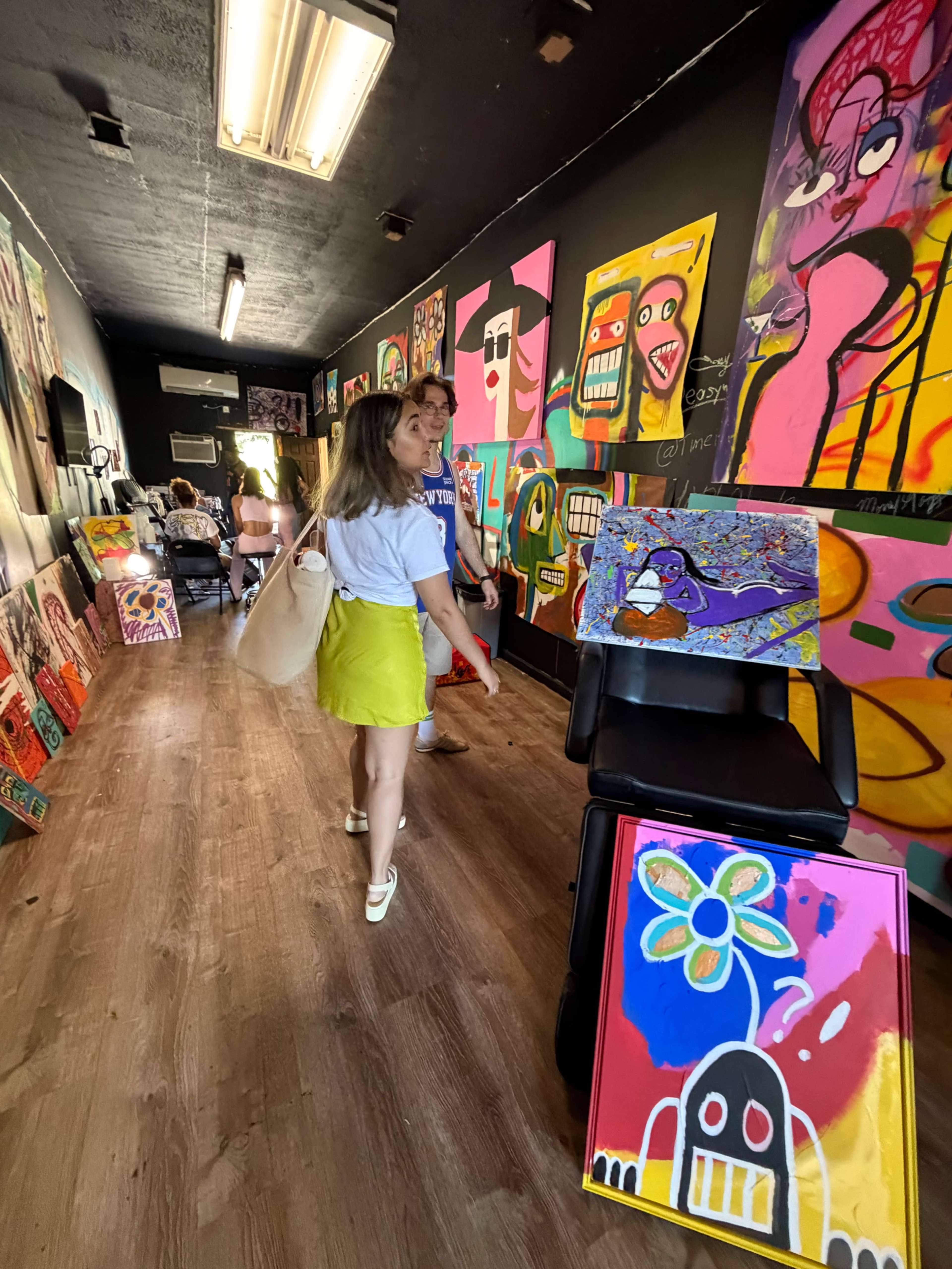 Two individuals walk through a vivid art gallery decorated with colorful paintings on the walls and on display stands.
