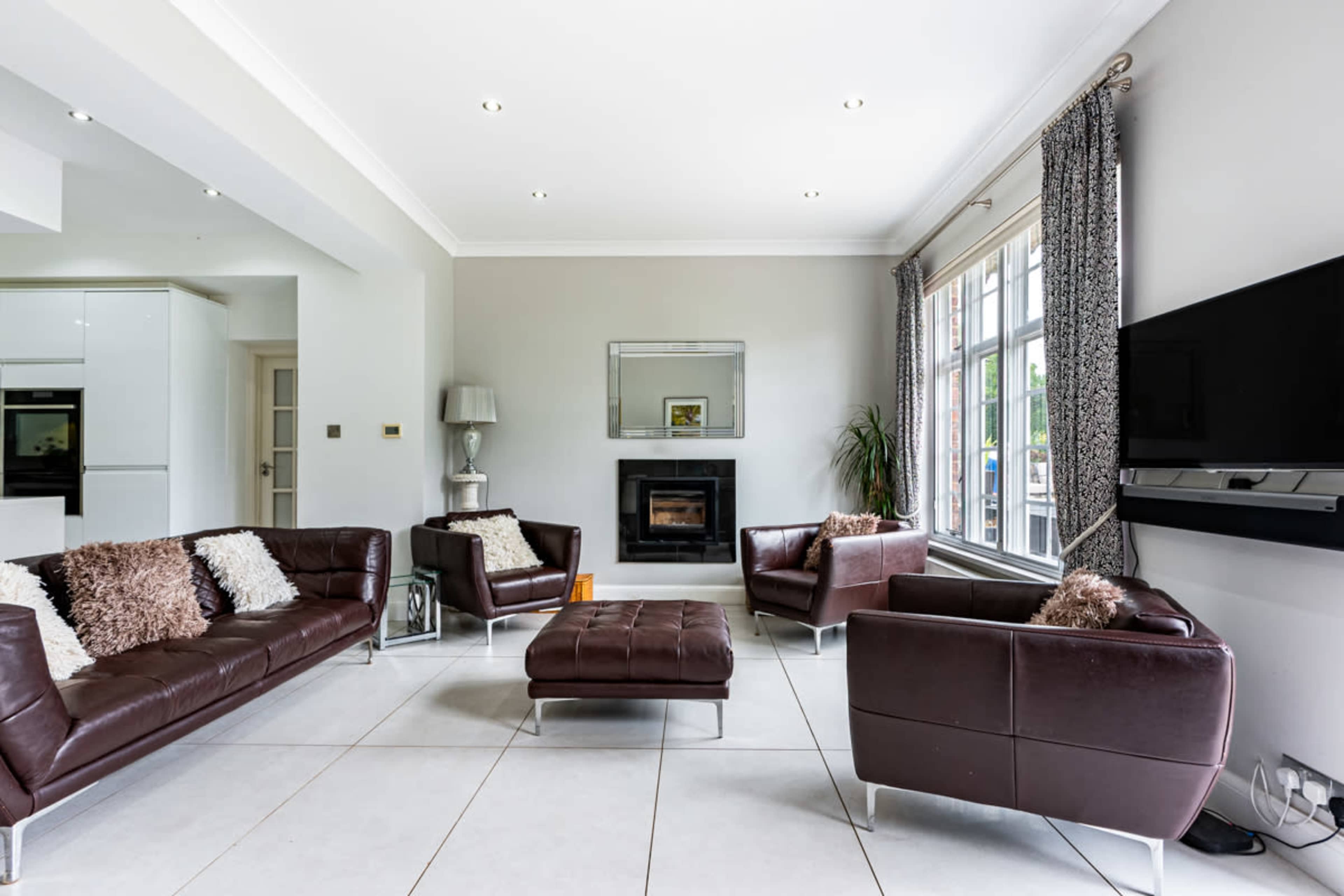 A modern living room featuring brown leather sofas, a fireplace, large windows, and a flat-screen TV mounted on the wall.