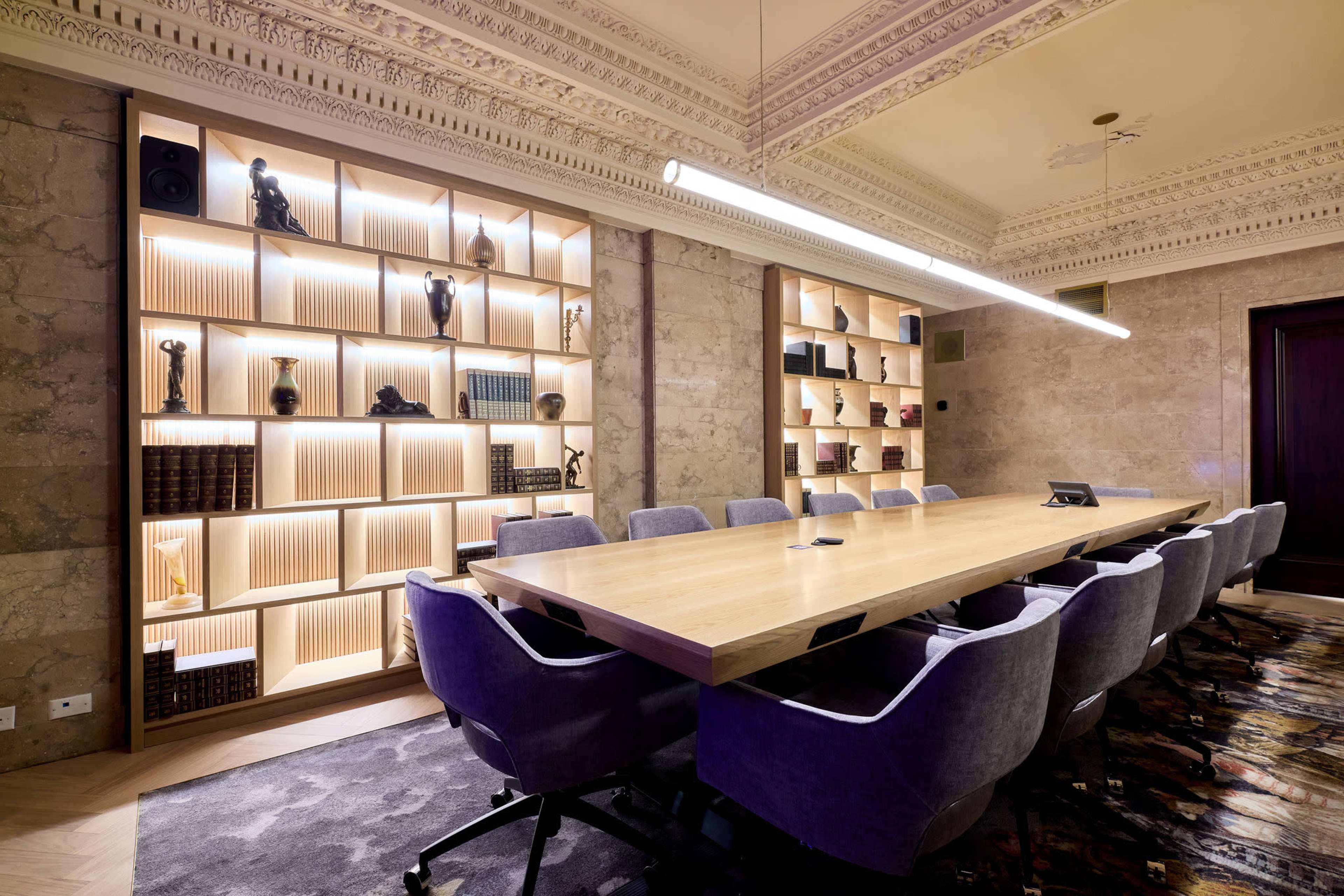 The image shows a modern conference room featuring a long wooden table surrounded by upholstered chairs, with illuminated shelves displaying various decorative items in the background.