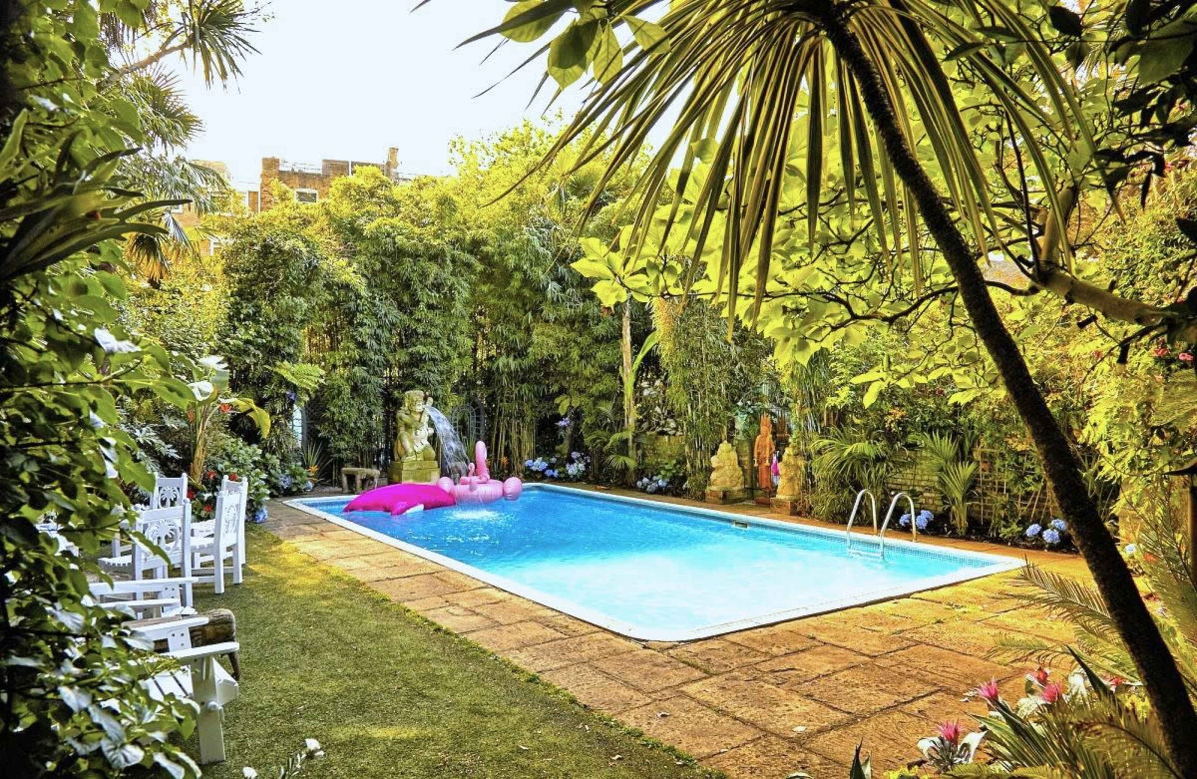 The image shows a private swimming pool surrounded by lush greenery and tropical plants, with pink floaties in the water and white chairs positioned nearby.