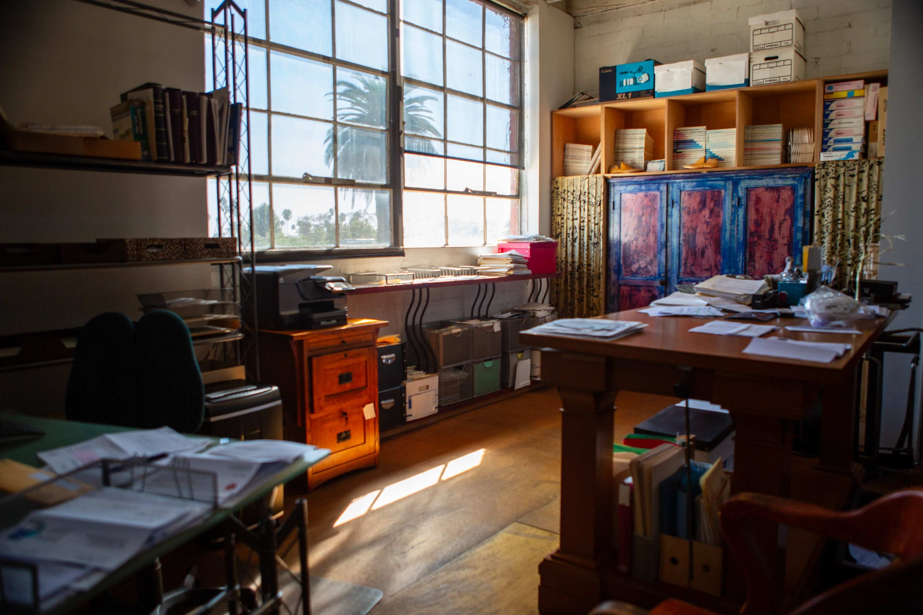 A bright office space features a large window, wooden furniture, and shelves filled with various documents and supplies.